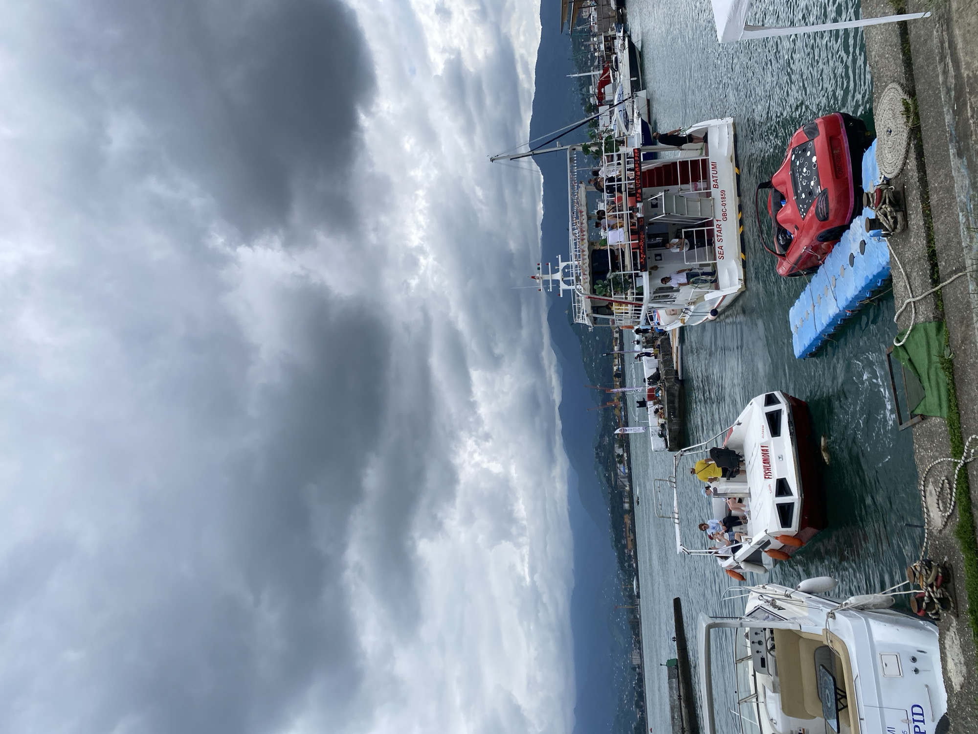 In Batumi, Georgia, a cloudy day casts a somber mood over a harbor scene. Several boats are docked, including a larger passenger vessel named "SEA STAR 1 BATUMI" with people on its deck and a smaller boat labeled "FISHLANDIA 1." A unique red convertible car is partially submerged next to a blue floating dock. The background shows a coastal city with mountains in the distance.