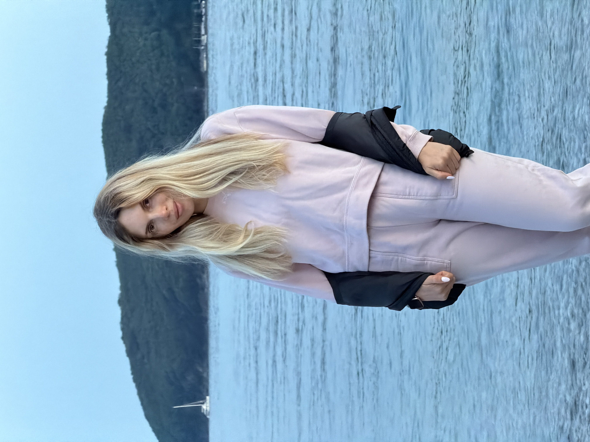 A young woman with long blonde hair is standing against the backdrop of the Adriatic Sea in Budva, Montenegro. She is wearing a light pink sweat suit and a black jacket is slung over her shoulders. She is looking at the camera. The background consists of the sea and a green hillside or island, with a small sailboat visible in the distance. The overall atmosphere appears calm and overcast.