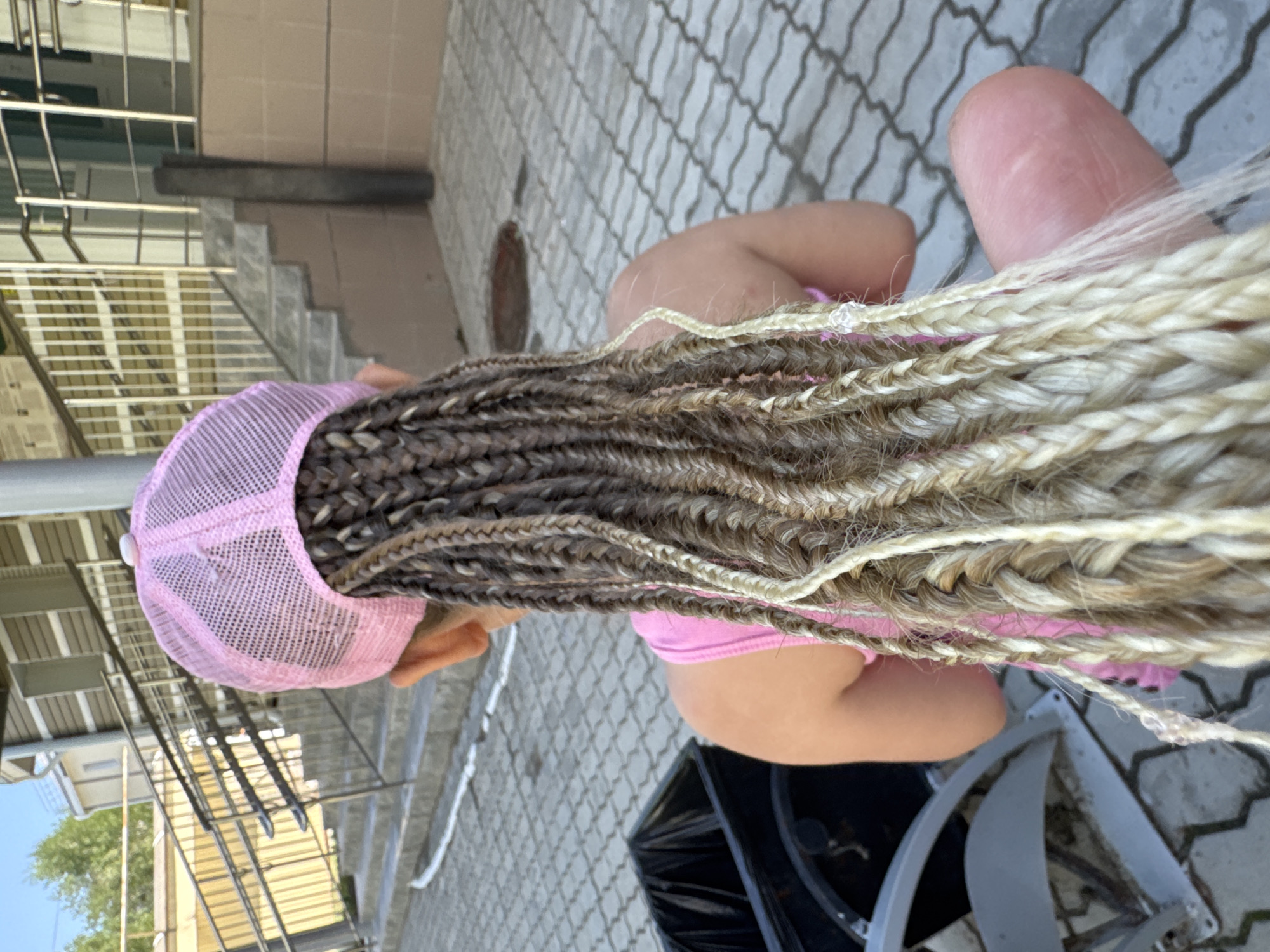 A young person wearing a pink mesh baseball cap and a pink tank top is seen from behind. Their hair is styled in numerous long, thin braids that extend down their back. Some braids are dark brown, while others are a lighter blonde, with some strands appearing to be lighter still. The person is sitting on the ground, which is paved with gray interlocking hexagonal tiles. In the foreground to the left, a black trash receptacle with a silver rim is partially visible. In the background, there is a set of outdoor stairs with metal railings and a building with light-colored siding and windows. To the far left, some green foliage and a blue sky are visible. The overall setting appears to be outdoors, possibly in a public area or courtyard within the city of Megion, Russia, given the architectural style and paving.