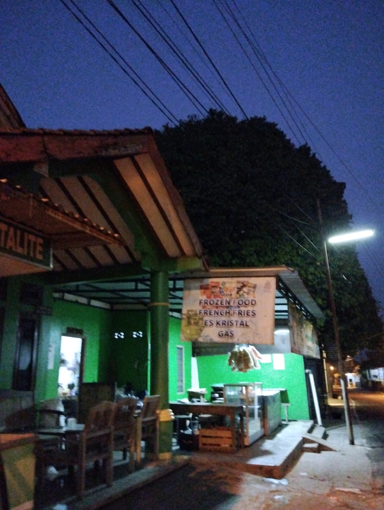 The image depicts a street scene at dusk or night in Salatiga, Indonesia, likely in front of a small food establishment. Overhead, power lines are visible against a dark blue sky. A building with a green wall and a brown and yellow striped awning or roof overhang is prominent. A sign attached to the awning reads "FROZEN FOOD FRENCH FRIES ES KRISTAL GAS". Beneath the awning, a wooden table and chairs are set up. Further to the right, a street lamp illuminates the area, casting shadows on the ground. In the background, trees and other structures are dimly visible. The overall impression is that of a modest, open-air eatery or street vendor.