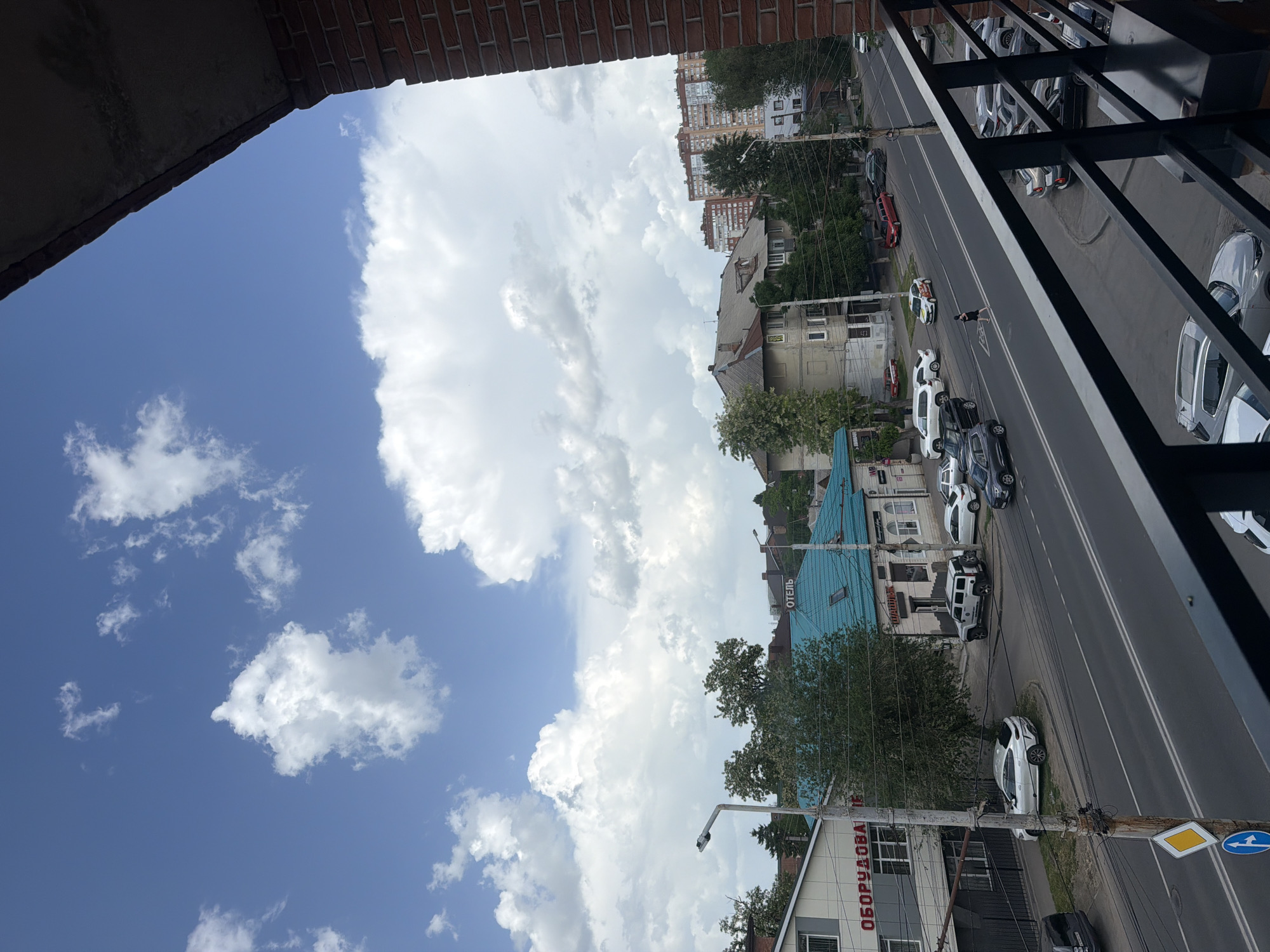 The image is a street view taken in Rostov-on-Don, Russia, partially framed by a brick building element on the left and a metal railing on the right. The street features parked cars on both sides and moving traffic. In the foreground, there is a utility pole with signage near a building that has "ОБОРУДОВА" written on it in Cyrillic. Further down the street is a building with a blue roof and a sign that reads "ОТЕЛЬ" (Hotel). A pedestrian is crossing the street. Above the buildings, there is a view of the sky with a mix of blue and cumulus clouds.