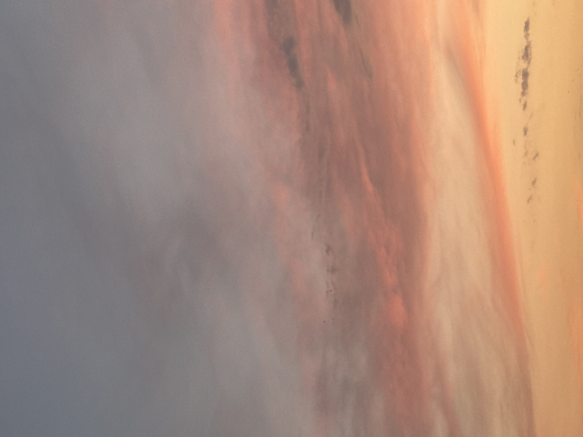 The image captures a sky filled with wispy clouds during either sunrise or sunset. The colors range from a soft gray at the top, transitioning through various shades of pink and orange towards the bottom. The lighting suggests it is either early morning or late evening. There are no discernable landmarks or city elements visible, making it impossible to identify a specific location within Bishkek City, Kyrgyzstan. The scene is entirely natural, focusing on the atmospheric conditions.