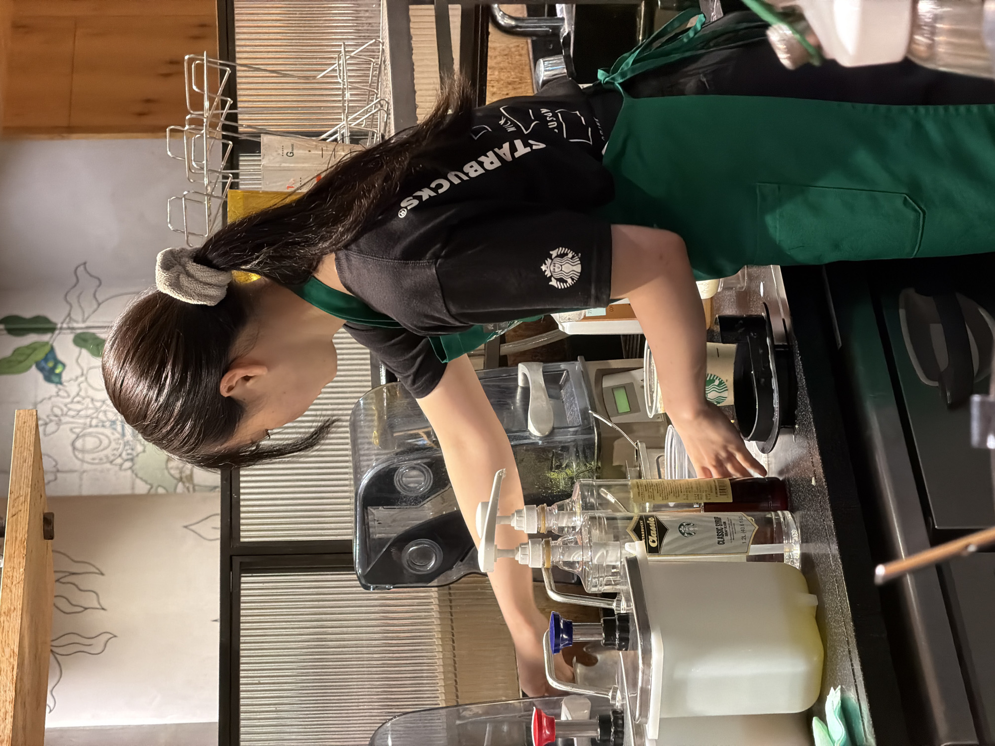 A young woman with dark hair tied in a ponytail is preparing drinks behind a counter at a Starbucks in Chuo, Japan. She is wearing a black Starbucks t-shirt and a green apron. In front of her are various drink preparation items, including syrup bottles, dispensers, and cups. Behind her, a drink machine is visible, as well as shelves with coffee-related supplies. The background shows a wall with a decorative mural.