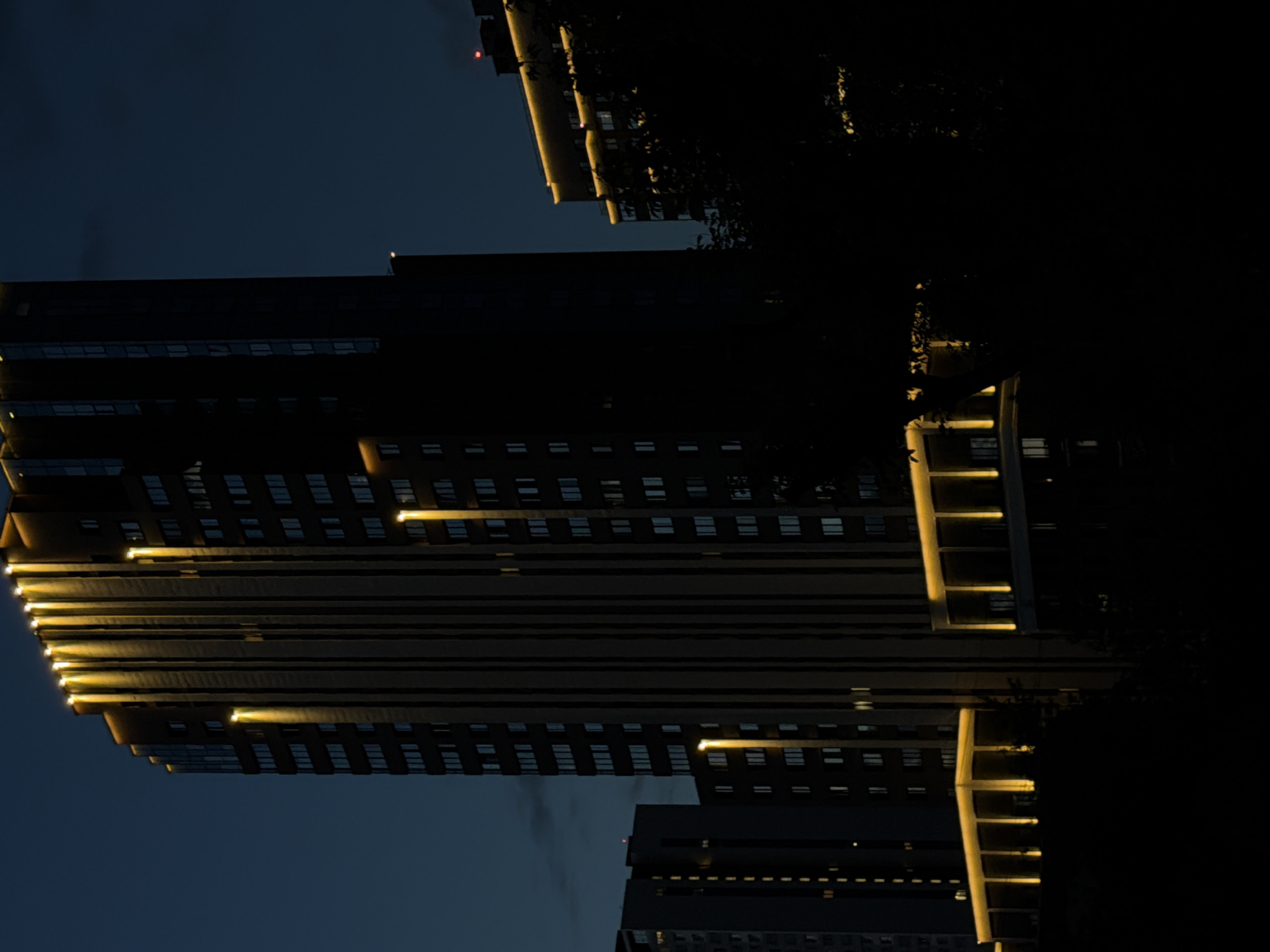 This image depicts a night scene of modern, high-rise residential buildings in Yekaterinburg, Russia. The primary focus is a tall, dark building with vertical architectural elements illuminated by warm, yellow uplighting along its edges and interspersed vertical strips. To the left, a slightly shorter, darker building with visible windows is partially obscured by the main structure. In the background and to the right, another building is visible, also illuminated by a band of warm light along its upper edge. The sky is a deep, dark blue, suggesting twilight or night. The foreground is dominated by dark shadows, with hints of foliage visible, suggesting trees or bushes. There are no people or visible actions depicted. The overall impression is one of urban architecture at night, with an emphasis on artificial lighting accentuating the structures against the dark sky.