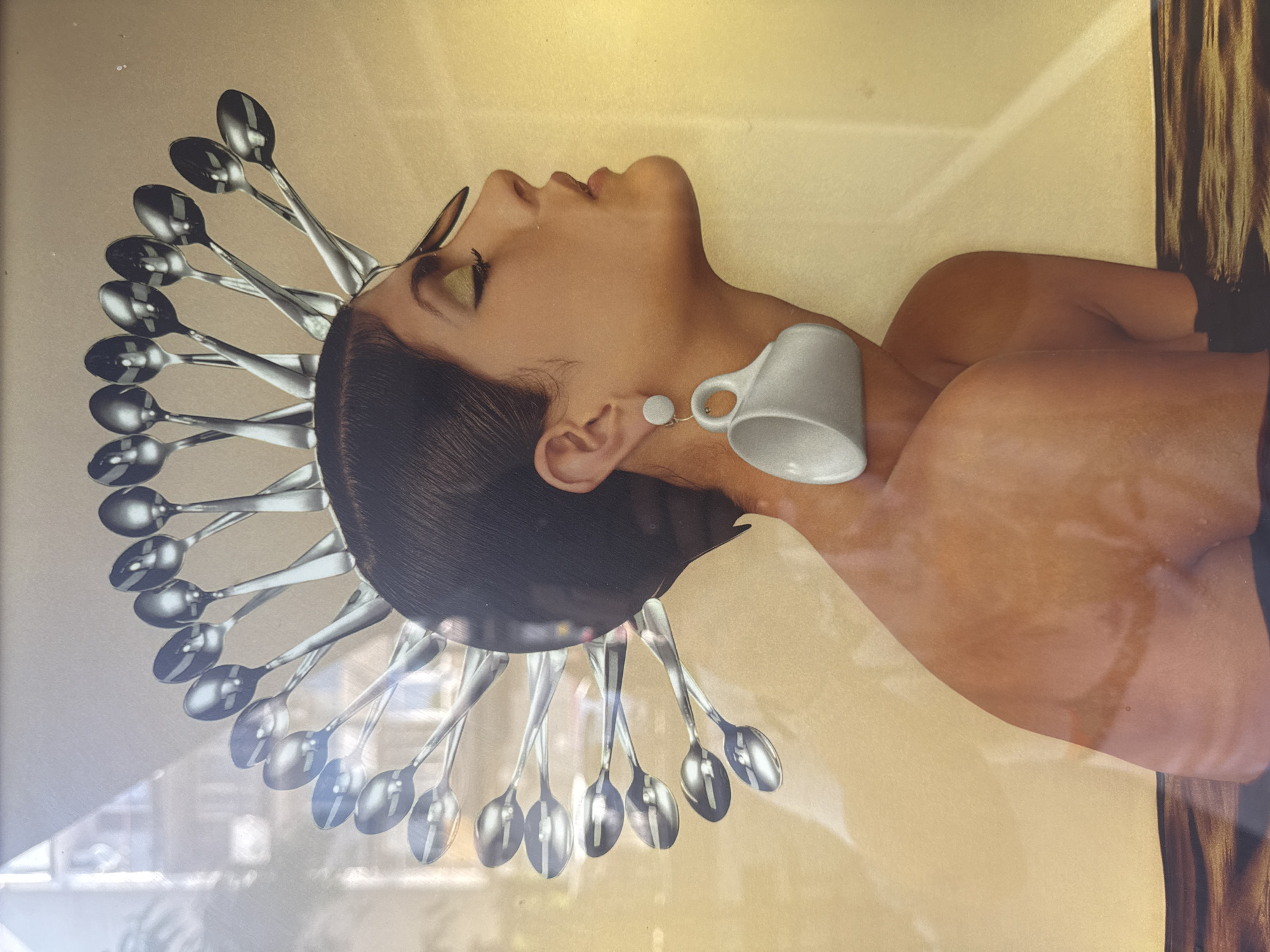 The image shows a framed piece of collage art. The artwork is dominated by a profile of a light-skinned woman with dark hair. Her face is turned upwards, and she wears golden eyeshadow. Around her head are arranged silver spoons radiating outward like a halo or headdress. A white coffee cup is attached to her neck as an earring. The background is a pale yellow or beige color. The frame is partially visible, showing a brown, textured edge on the right side. The picture was taken in Plovdiv, Bulgaria.