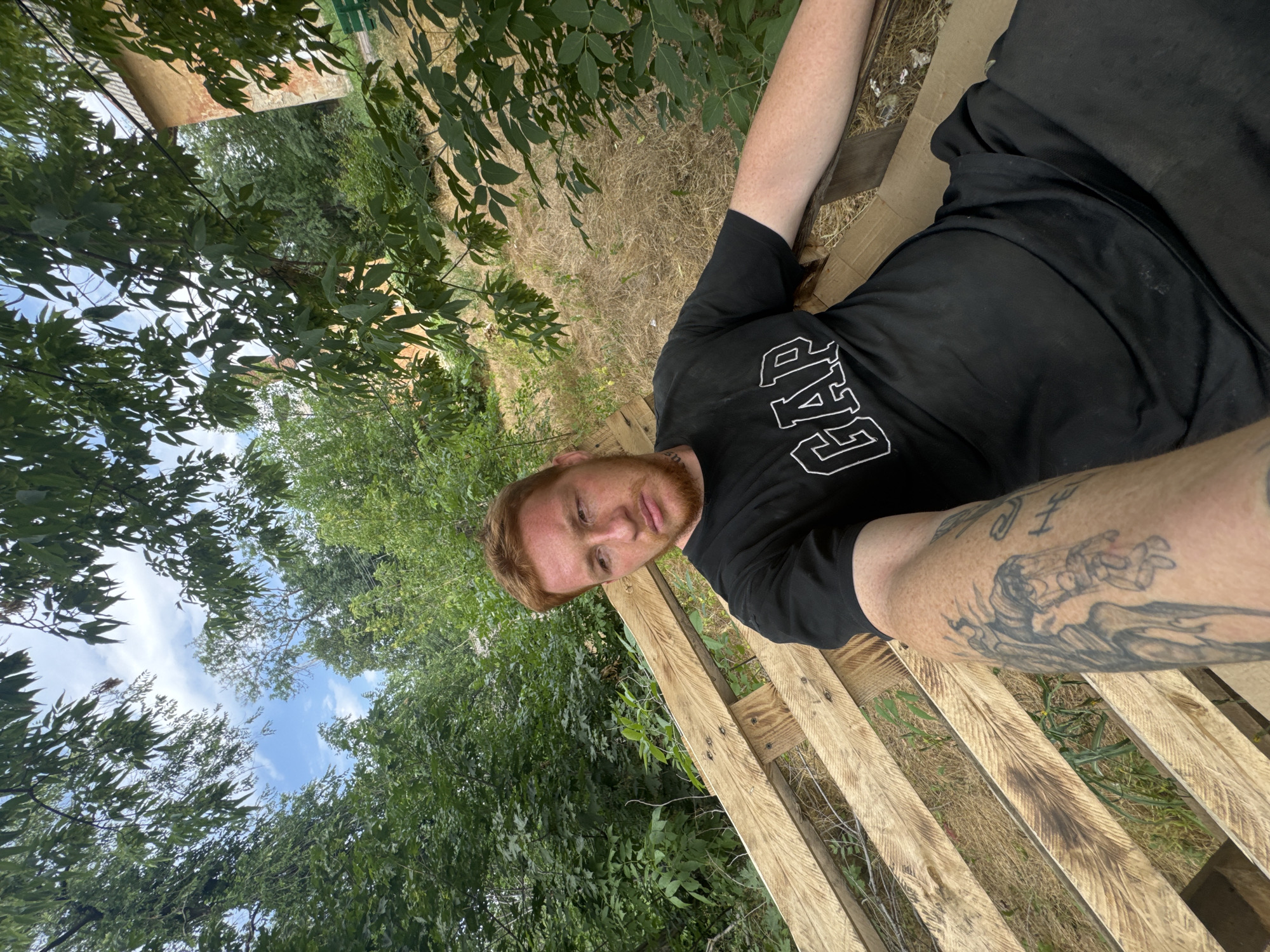 A man with ginger hair and a beard is sitting on a wooden pallet bench outdoors. He is wearing a black t-shirt with a white "GAP" logo and black pants. His left arm, which is visible in the foreground, has tattoos. The man is looking towards the camera. The background consists of trees, dry grass, and a brick building. The sky is visible through the trees. The scene appears to be in a rural or semi-rural setting.