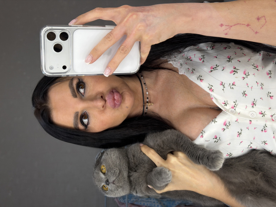 An adult female, identified as being in Svkh Voronezhskiy, Russia, is depicted in an indoor setting with a plain dark grey background. She has long dark hair, prominent eyebrows, and significant eye makeup. Her lips are pursed in a "kissing" expression, and her gaze is directed towards the smartphone she holds in her left hand, indicating she is taking a selfie. The phone is light-colored, housed in a clear case, and features a triple-lens camera array on its rear. Her fingernails display a French manicure. She wears a white V-neck top with pink floral patterns and lace trim, revealing cleavage. A black choker necklace with beads and a small golden cross pendant is visible around her neck. On her left forearm, a faded pink tattoo resembling a constellation is present. She cradles a grey Scottish Fold cat in her right arm. The cat has amber eyes and is looking upwards and to its right.