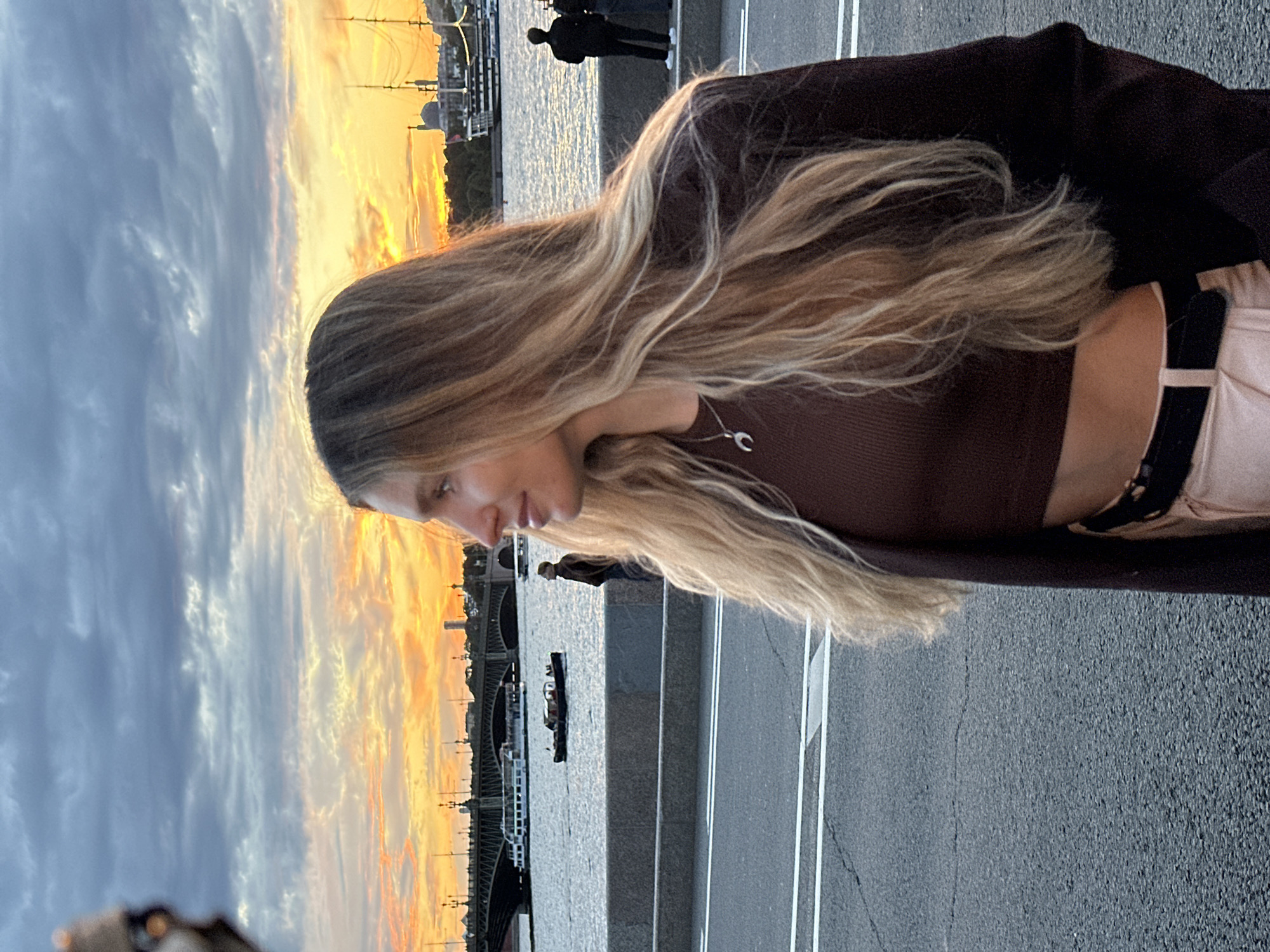 A medium shot captures a young woman from the chest up, facing right, with long blonde hair flowing over her shoulders. She wears a dark brown fitted top and a dark brown jacket, and a silver crescent moon pendant necklace. She is on a paved road with white lines, with a stone embankment to her left. Beyond the embankment is a body of water, likely the Neva River, with boats and a bridge in the distance. The sky is filled with clouds and a vibrant sunset in shades of orange and yellow. In the background, on the riverbank, are buildings and two figures standing. This scene is consistent with the embankments of the Neva River in Saint Petersburg, Russia, particularly near one of its many bridges, during sunset.