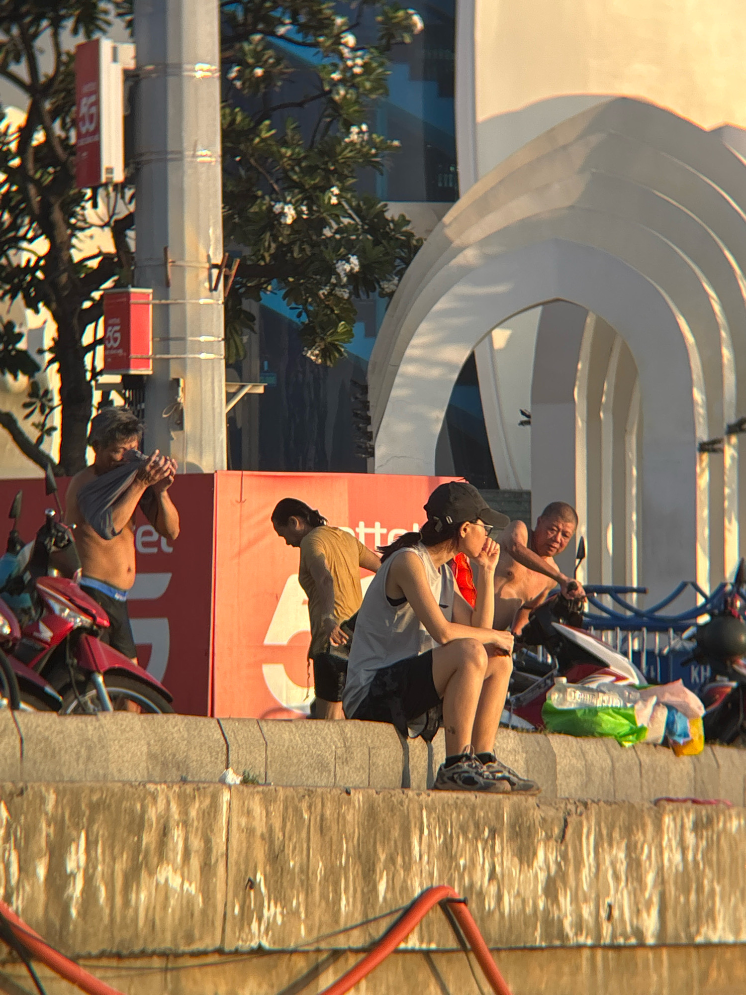 The image depicts a group of individuals situated along a concrete barrier in what appears to be Nha Trang, Vietnam. Sunlight illuminates the scene, casting long shadows and highlighting the textures of the environment.
In the foreground, to the left, a man is seen partially disrobing, holding a grey t-shirt in his hands. He is wearing dark shorts. Adjacent to him, a red motorcycle is parked. Further to the right, another man in a mustard-colored t-shirt is bent over.
In the middle ground, a person wearing a black baseball cap and glasses sits on the concrete barrier, facing left, with their hand near their mouth. They are wearing a grey sleeveless top and dark shorts. To their right, another man, shirtless, is leaning forward, appearing to interact with a motorcycle parked behind the barrier.
Behind the individuals, a vibrant red wall with white lettering, possibly advertising "5G", is visible. In the background, architectural elements with arched structures can be seen, illuminated by the warm light. Trees with foliage and white flowers are also present, adding to the urban landscape. The overall impression is of a casual gathering in a public space, likely a promenade or waterfront area in Nha Trang.