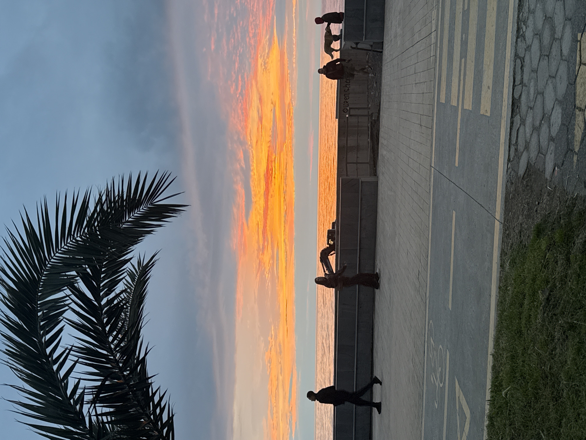 The image shows a sunset view from a location in Batumi, Georgia. The sky features a vibrant orange and yellow sunset with some cloudy areas. There's a palm tree in the left foreground. A paved walkway is visible with markings indicating a bicycle lane. There are several people present along the walkway, some standing and interacting near a short wall overlooking the sea. A lone individual is walking along the walkway. The water reflects some of the sunset's colors.