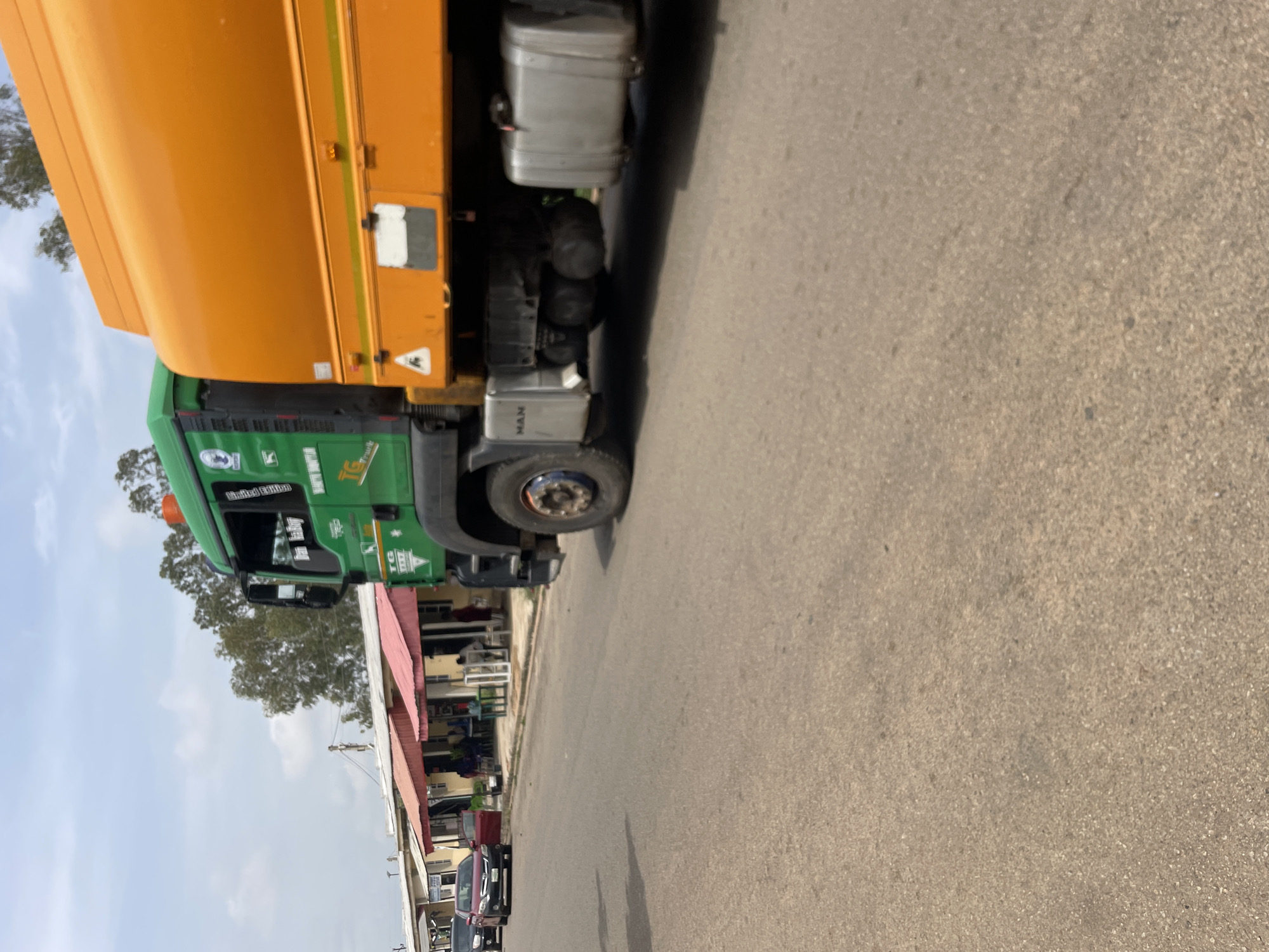 A large green and orange truck is parked on the side of a paved road in the city of Jos, Nigeria. The truck has an orange tank on the back and a green cab. The word "MAN" is visible on the side of the truck. In the background, there are several small buildings with red roofs and a few trees. There are also a few cars parked on the side of the road, and some people can be seen near the buildings. The sky is cloudy. The road appears to be in a residential or commercial area.