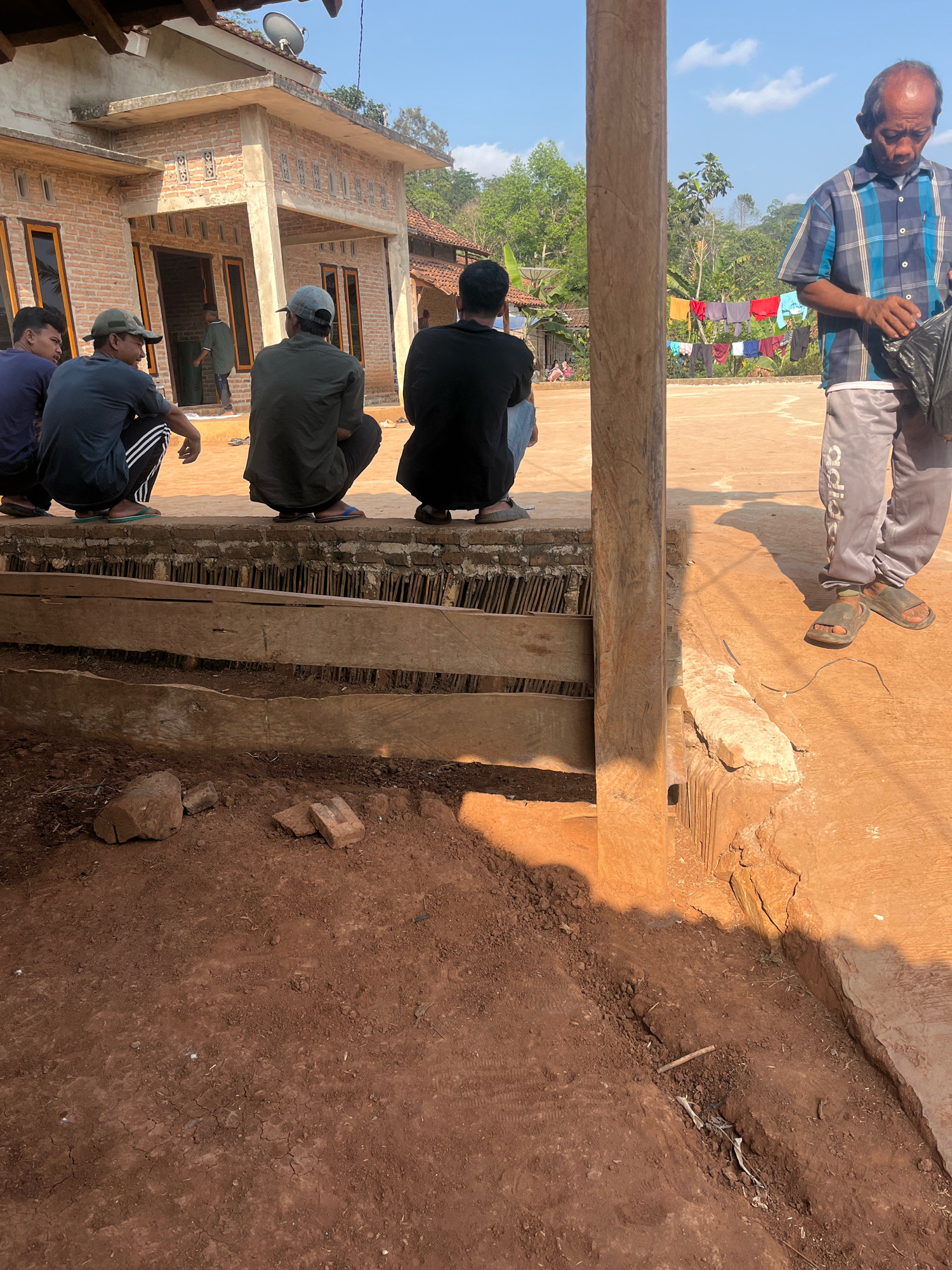 In Sidomulyo, Indonesia, a group of men are gathered outdoors in a rural setting. The scene is dominated by a low, brick building with an exposed concrete roof. In front of the building, several men are sitting or squatting on a low wall constructed with bricks and wooden planks. One man in the foreground, wearing a blue and white plaid shirt and grey sweatpants with "adidas" printed on them, is holding a black plastic bag. He is wearing dark sandals. To his left, four men are positioned on the wall, facing away from the camera. They are dressed casually in t-shirts and hats. One man wears a dark blue t-shirt, another a dark grey t-shirt and a green cap, a third a dark green t-shirt, and the fourth a black t-shirt. The ground in front of the wall is reddish-brown dirt, with some scattered bricks and debris. In the background, laundry is hanging on a clothesline between trees, and other buildings and lush vegetation are visible, indicating a residential or village area. The sky is clear and blue.