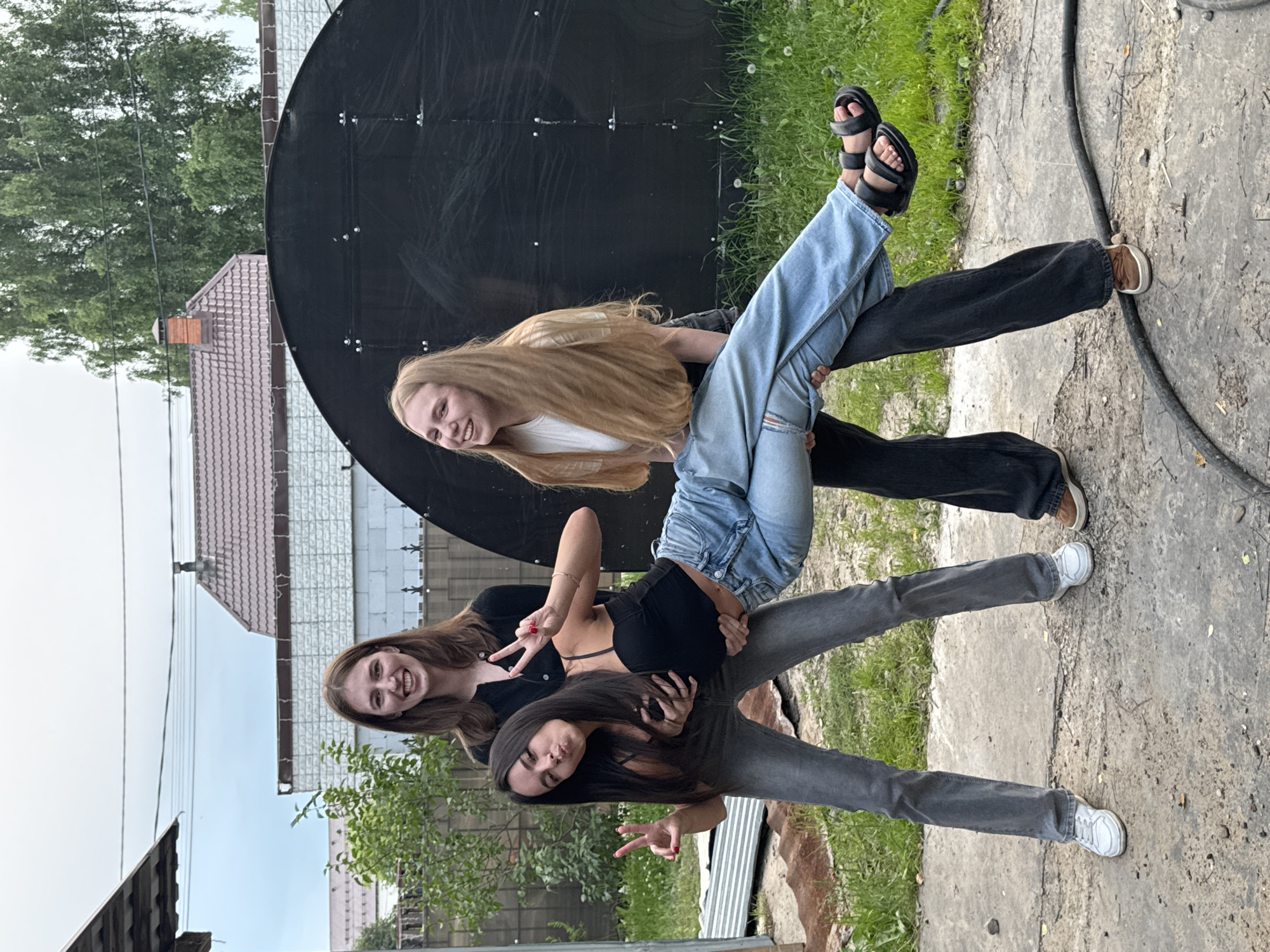 Three young women are posing for a photo outdoors. The woman in the center is being held horizontally by the two women on either side of her. She is wearing a black crop top and light blue jeans, and her arms are extended, holding up peace signs. The woman on the left is wearing grey jeans and a black cardigan, and she is also holding up a peace sign. The woman on the right is wearing a white t-shirt and dark pants, and she is smiling and looking at the camera. They are standing on a concrete surface in what appears to be a backyard or a rural area, with grass and some overgrown vegetation visible. In the background, there is a black, curved structure, a wooden fence, and houses with tiled roofs. The sky is overcast. The overall impression is a candid and playful moment captured between friends. The image does not contain any elements that would suggest a specific location within the city of Perm, Russia.