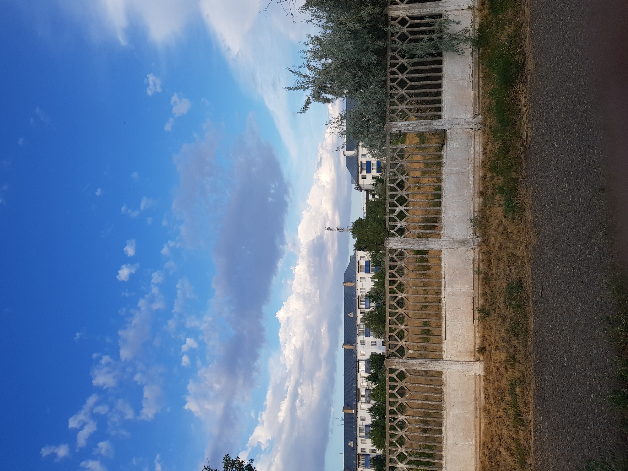 The image depicts an urban landscape in Kurchatov, Kazakhstan. In the foreground, a weathered concrete fence with a decorative diamond pattern runs horizontally across the frame. Behind the fence, dry, yellowed grass covers the ground, interspersed with patches of green vegetation. Beyond the grass is a row of multi-story apartment buildings with white facades, blue roofs, and blue window frames. Some buildings feature triangular gables on their roofs. Trees and foliage are visible between the buildings and in the background. The sky above is a vibrant blue, dotted with scattered white and gray clouds. A telecommunications tower is visible in the distance on the left side of the image, and a large, leafy tree with silvery-green foliage is prominent on the right side. The overall scene suggests a residential area under a bright, partly cloudy sky.