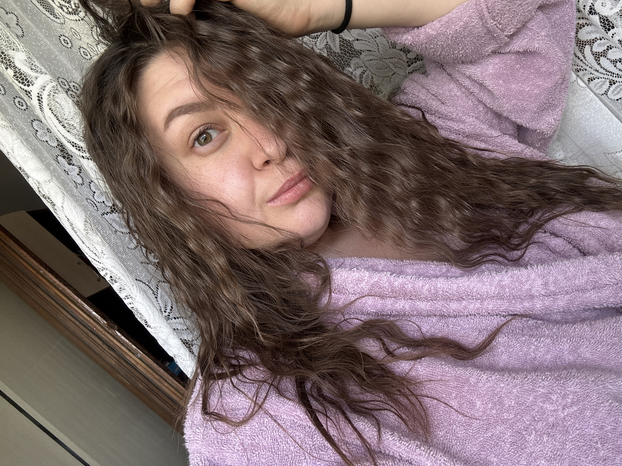 A woman with long, wavy brown hair partially covering her face is lying down, wearing a light purple terry cloth robe. Her left hand is raised, touching her hair near her scalp. The background includes a window with a white lace curtain and a dark frame. The location cannot be determined from the image.