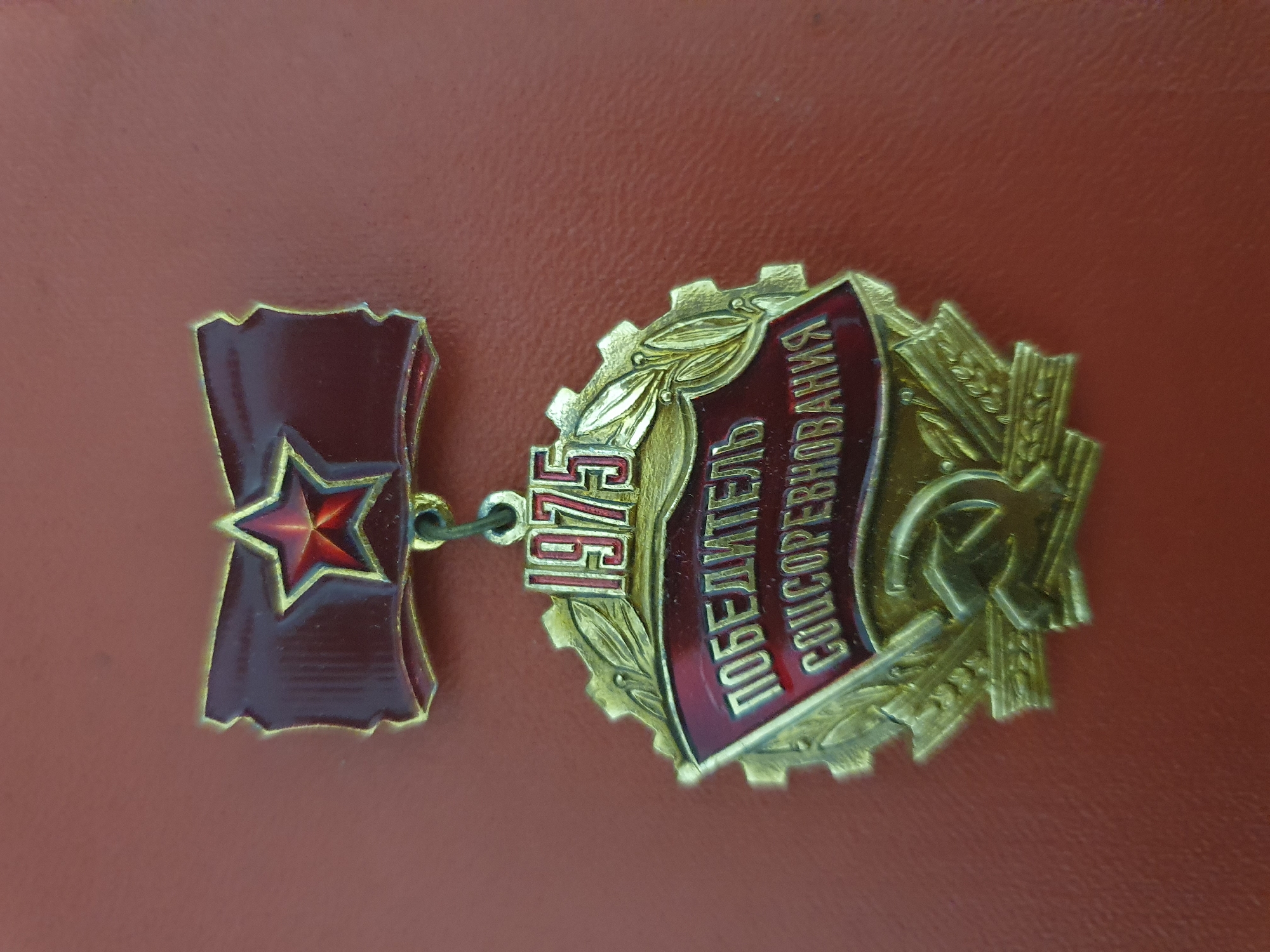 The image shows a close-up, top-down view of a Soviet-era medal for "Winner of Socialist Competition" from 1975. The medal is gold-colored with red enamel accents. It features a red star on a ribbon at the top, the year "1975" in red numerals, a banner with the inscription "ПОБЕДИТЕЛЬ СОЦСОРЕВНОВАНИЯ" (Winner of Socialist Competition) in white Cyrillic letters, and a hammer and sickle emblem at the bottom, all surrounded by laurel branches and a cogwheel. The medal is displayed against a textured, reddish-brown surface, possibly a leather-bound album or display case. There are no people, actions, or interactions visible in the image. The location is not specified in the image.