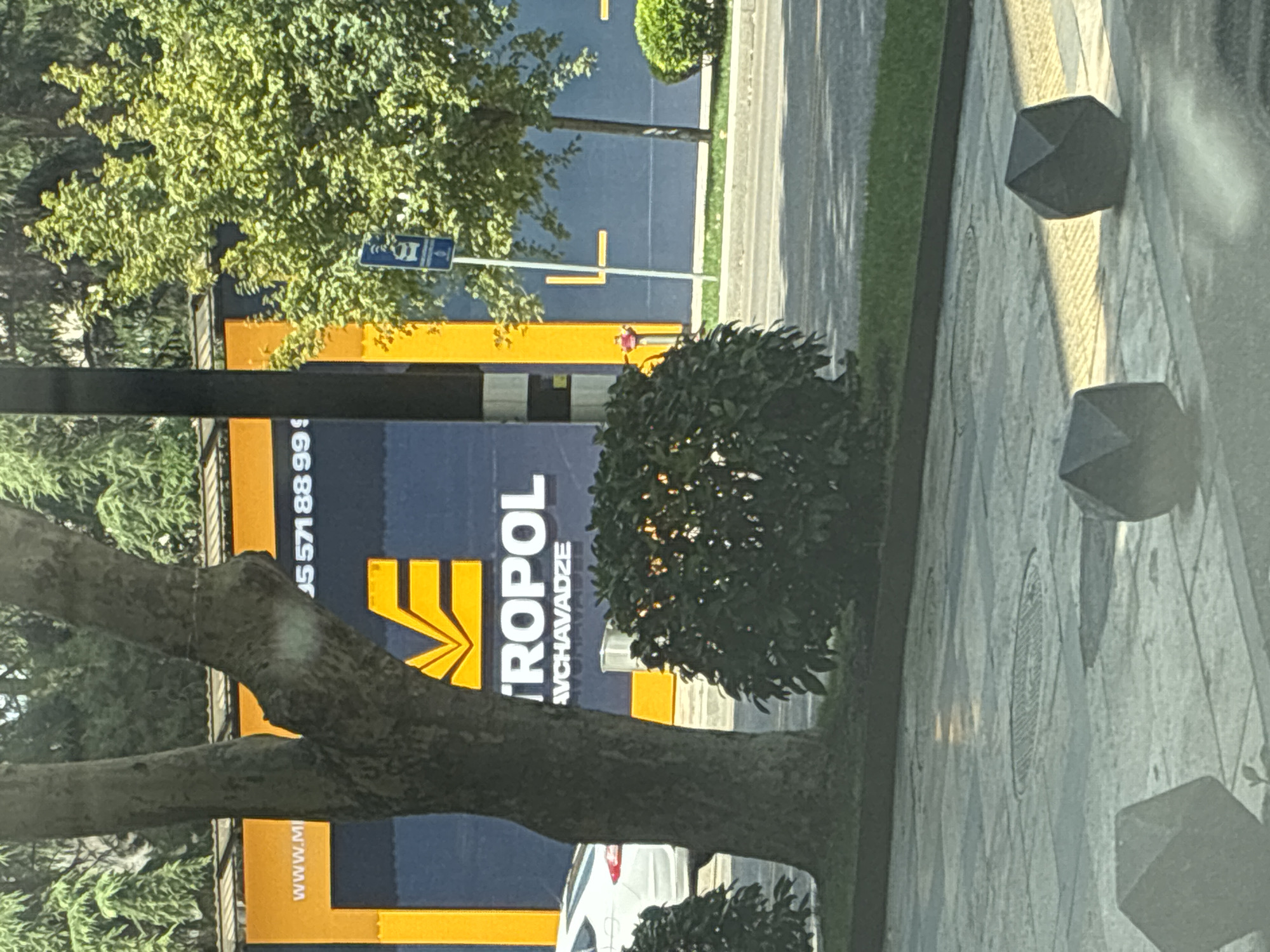 The image captures a view of a building facade with a large blue and yellow advertisement. The text on the advertisement reads "WWW.M", followed by what appears to be a phone number starting with "+95 571 88 99", and "METROPOL AVCHAVADZE". The advertisement features a prominent yellow geometric logo. A tree trunk partially obscures the left side of the advertisement. To the right of the tree, a portion of a white car is visible. In the background, there are green trees and foliage. A blue street sign with a symbol of a bus is visible on a pole. The foreground shows a paved area with geometric bollards, and a section of a street with green grass. The overall scene suggests an urban environment in Tbilisi, Georgia, likely near a street or commercial area.