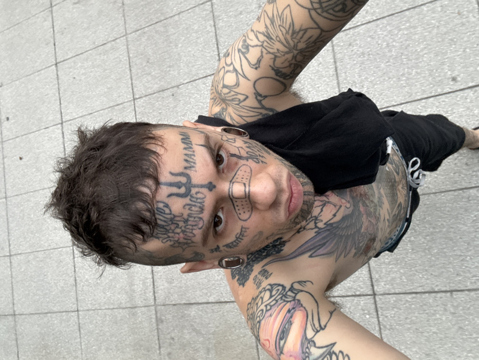 A person with extensive tattoos is looking up at the camera. The tattoos cover their face, neck, chest, and arms. On their face are the words "MAMMA" and religious symbols; a bandage is on their nose. They have piercings in their earlobes. They are wearing a black top and bottoms. The background is a tiled surface.