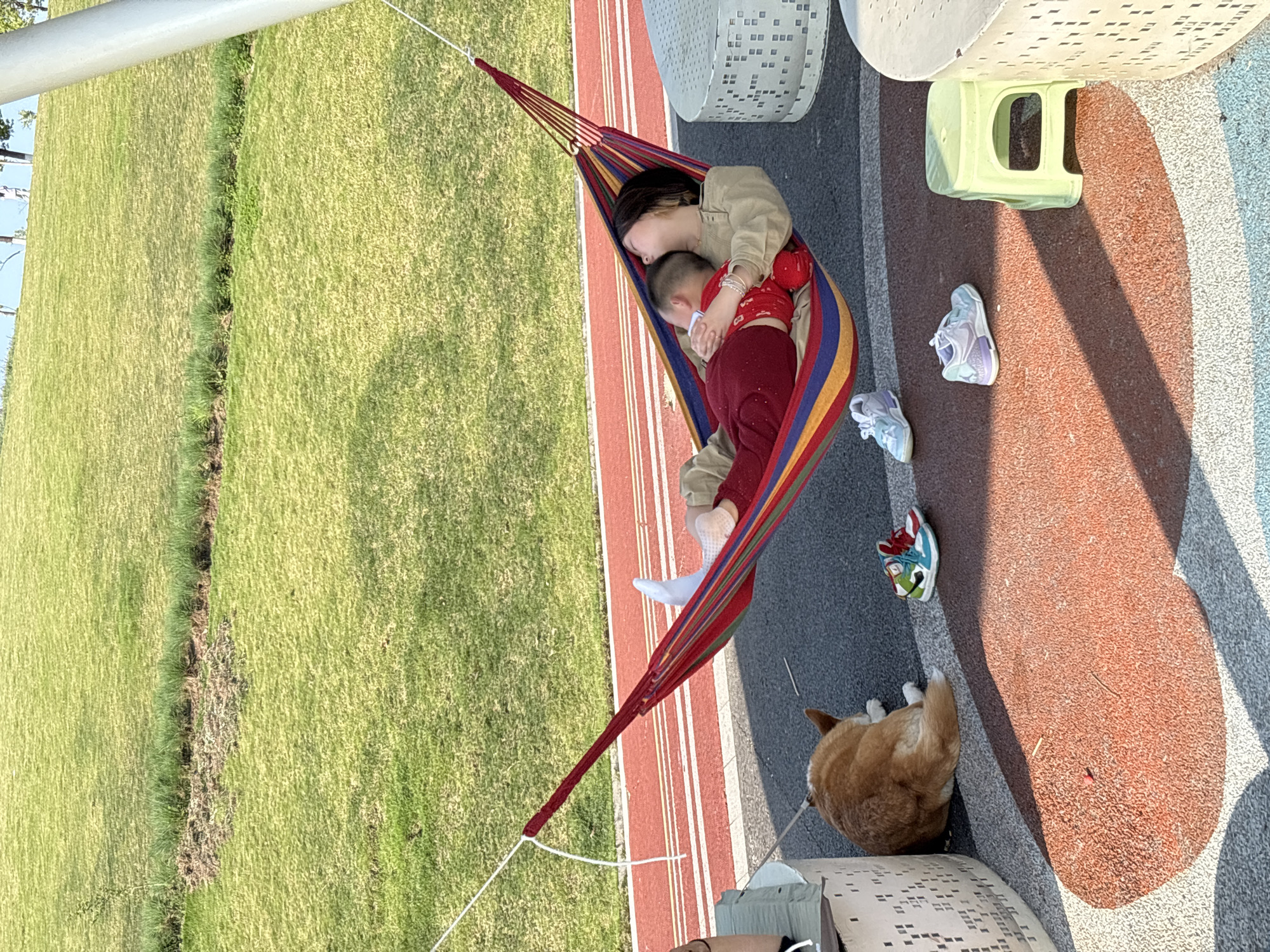 A photo taken in Jiangjin, China shows a woman and a child lying in a hammock slung between supports next to a grassy area. The woman is wearing a light jacket and the child is wearing red clothing. Several pairs of shoes are on the ground near the hammock, along with a dog. The ground is a mix of red, gray, and striped surfaces. A white metal structure with square cutouts is nearby.