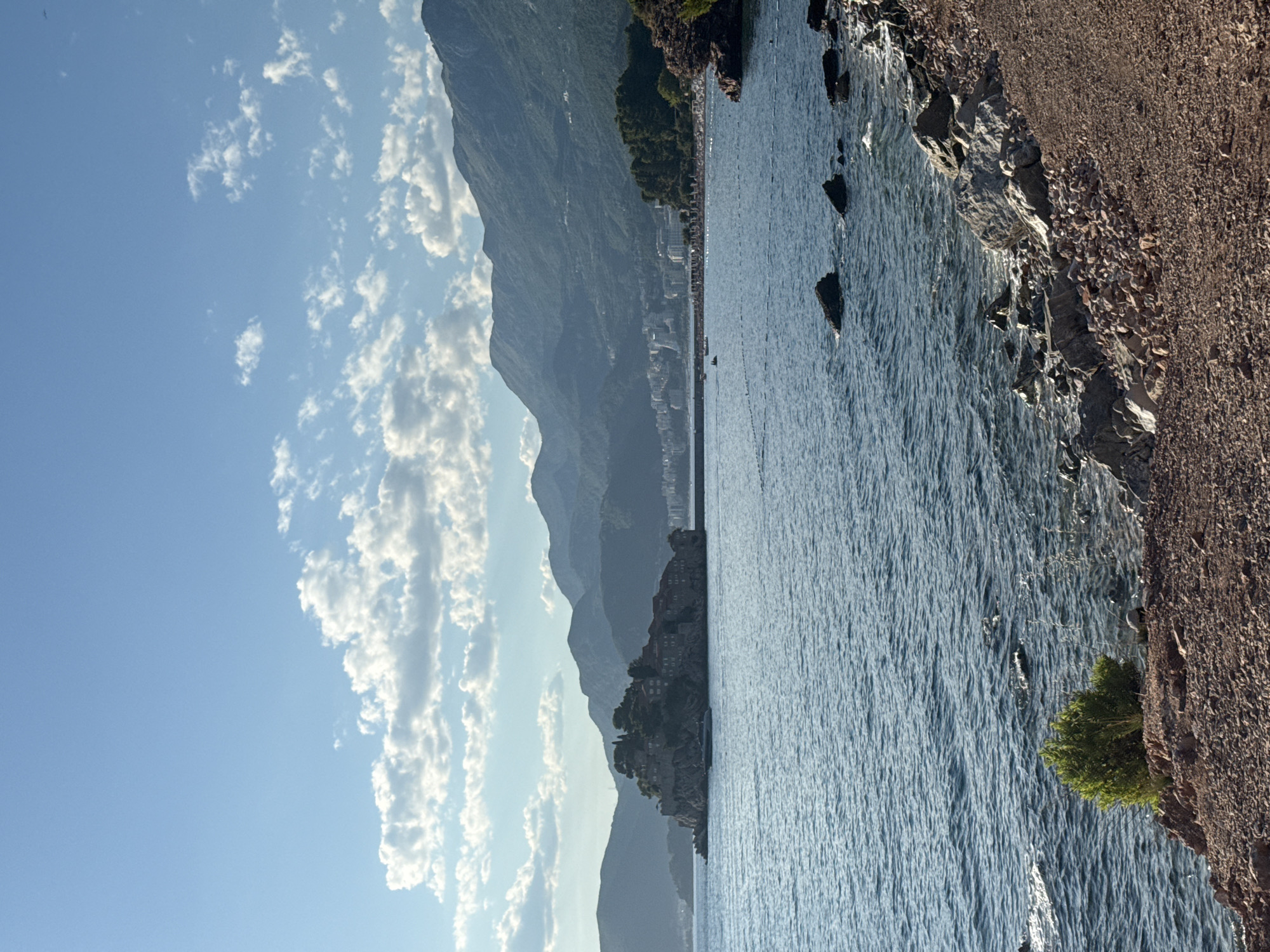 The image depicts a scenic coastal view in Sveti Stefan, Montenegro. In the foreground, a rocky shoreline is visible, with a small green shrub growing on the left side. The sea stretches out, with gentle waves and scattered rocks. In the middle ground, the picturesque island of Sveti Stefan, a fortified village with red-tiled roofs and stone buildings, is connected to the mainland by a narrow causeway. A sleek yacht is moored in the water near the island. Further in the distance, a city with a modern skyline of buildings lines the coast, backed by lush green hills and a chain of majestic mountains that fade into the hazy horizon. The sky above is a clear blue, dotted with scattered white clouds, suggesting a pleasant, sunny day.