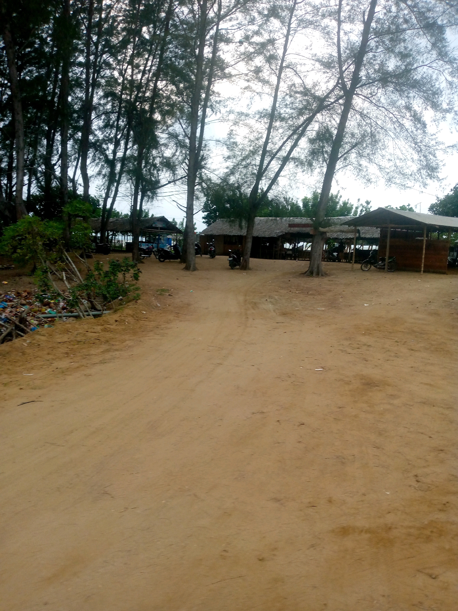 The image depicts an outdoor scene, likely a roadside or a small village area, in Kutaglumpang, Indonesia. The foreground is dominated by a dirt road, appearing dry and sandy. To the left, there is a pile of accumulated trash, consisting of plastic bottles, bags, and debris, situated near some vegetation and wooden structures. In the middle ground and background, there are several open-air shelters with thatched roofs, some of which appear to be small shops or food stalls. A few motorcycles are parked near these structures. Tall, slender trees with sparse foliage are scattered throughout the scene, their branches reaching towards an overcast sky. The overall impression is of a somewhat unkempt but functional area.