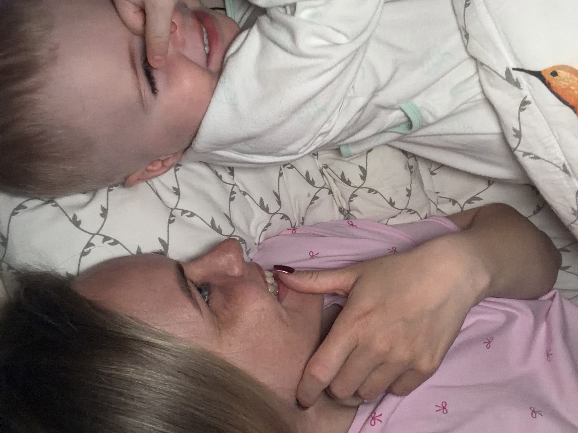 A woman and a baby are lying in bed. The woman is on her side, facing left, and the baby is above her, facing the top of the frame. The woman is wearing a pink top with bow decorations, and she has light brown hair. The baby is wearing a white onesie and has short blond hair. The sheets have a gray pattern. It is impossible to determine the location in the city of Votkinsk, Russia, from the provided image.