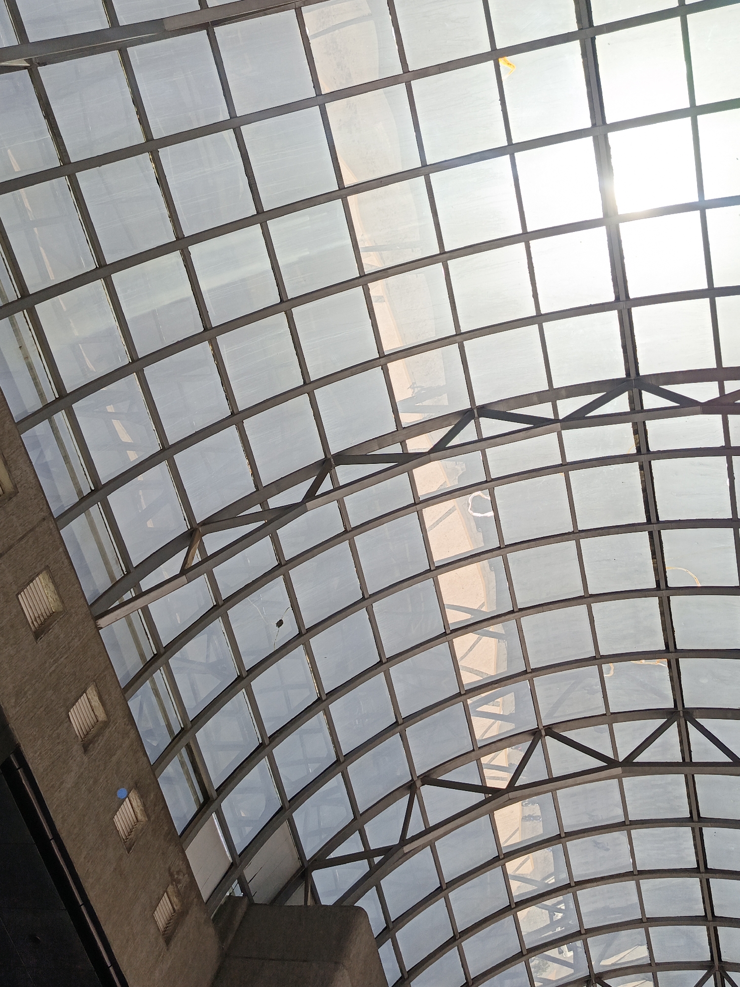 The image is an upward-angled view of a large, curved glass roof structure with a grid pattern. The structure appears to be composed of square or rectangular glass panels supported by a metal frame. Sunlight filters through the glass, creating bright patches and casting shadows. The roof is curved, suggesting a barrel vault or similar architectural design. A partial view of a building exterior is visible on the left side of the frame, revealing a brick or stone facade with small, rectangular window openings. The location is Unknown, Unknown. The scene is static, with no visible people or actions taking place.