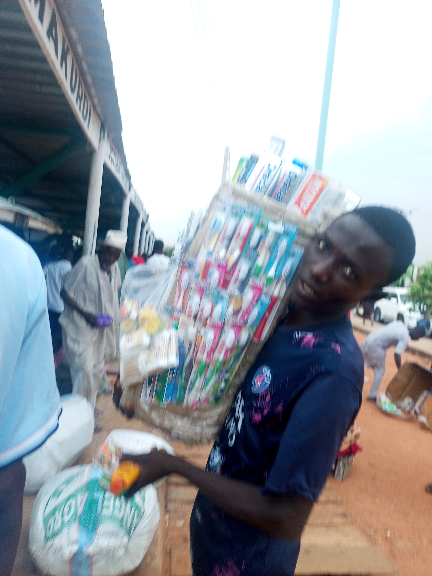 The image shows a young man in a navy blue Paris Saint-Germain t-shirt carrying a large bundle of toothbrushes on his back. He is looking at the camera with a slightly blurred background, which suggests he might be in motion. The location appears to be a bustling market area, possibly in Makurdi, as a sign with "MAKURDI" is partially visible in the background. Other people are visible in the background, including a man in a white robe and hat and another person bending over. Additional sacks are also visible, as well as cars in the distance. The environment appears to be outdoors on a partly cloudy day.