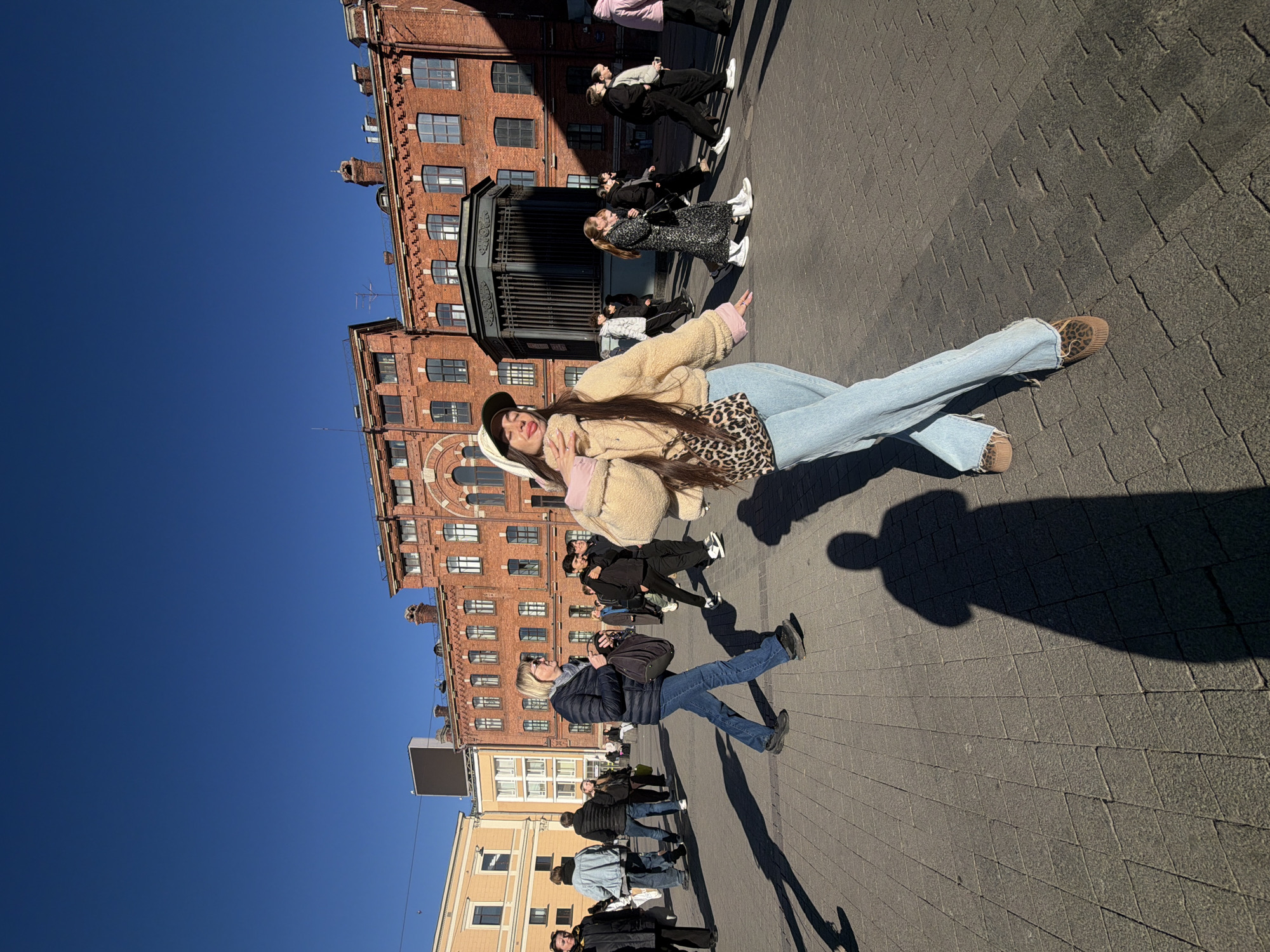 Here's a description of the image:
A woman is posing for a photo on a cobblestone street in what appears to be Saint Petersburg, Russia. She is wearing a beige sherpa jacket, blue jeans, and leopard print shoes. She is also wearing a hat. She is standing in front of a large brick building with many windows. The sky is blue. Other people are walking in the background.