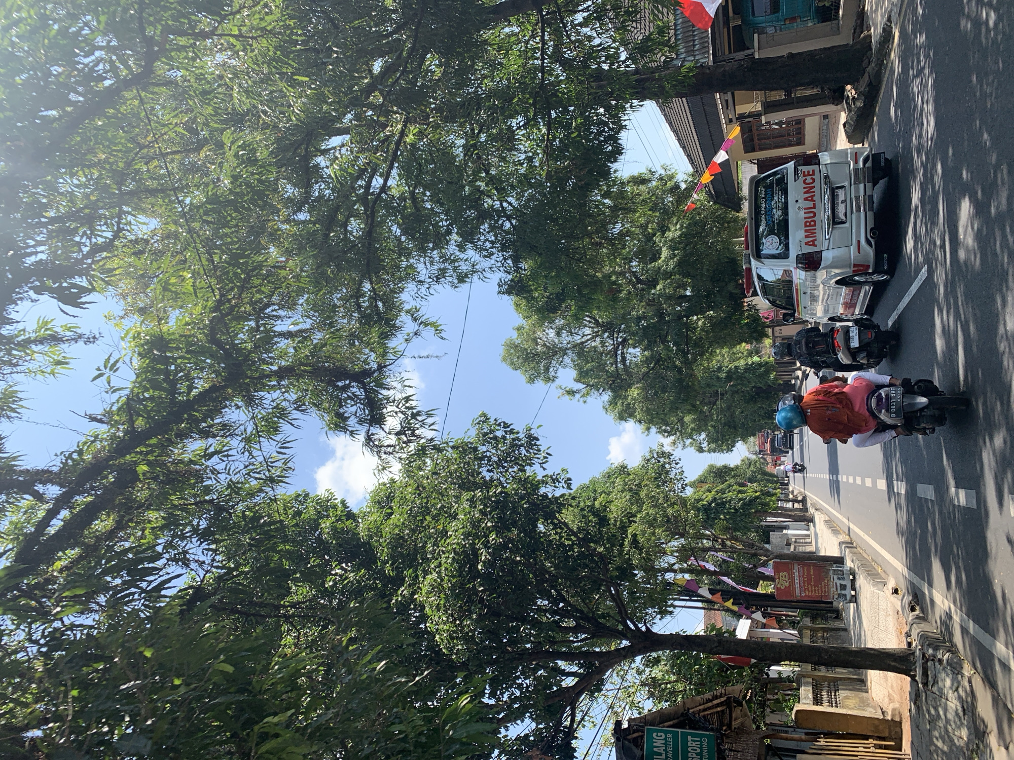 The image shows a sunny street scene in Ciamis, Indonesia, with lush green trees lining both sides of the road. A white ambulance with "AMBULANCE" written on its side is driving in the right lane, followed by a person on a motorcycle. In the left lane, another person is riding a motorcycle with a passenger wearing an orange shirt and a blue helmet. The road is paved, and there are houses and shops on either side. Flags are also visible, adding a festive touch to the scene. The sky is blue with a few white clouds.