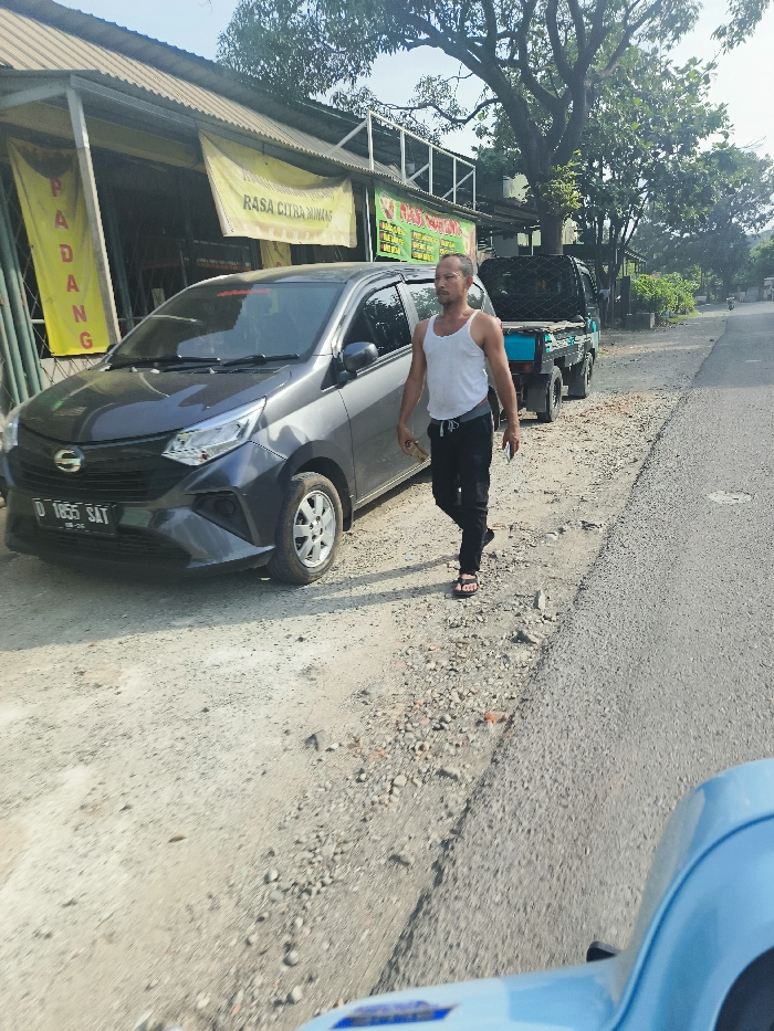 A man is walking on the right side of a paved road in front of a gray car. The car is parked on the left side of the road. The man is wearing a white tank top and black pants. He is walking away from the camera. The road is paved with asphalt. The man is walking on the shoulder of the road. There is a small building on the left side of the road, and a blue truck is parked in front of it. The building has a sign that says "RASA CITRA MINUM" in Indonesian. The man is looking at the ground as he walks. The scene is located in Jatiluhur, Indonesia (latitude: -6.578, longitude: 107.436). The sky is clear and the weather is sunny. The image was taken from a vehicle that is driving on the same road as the man. The photo was taken in the middle of the day, likely between 10 am and 4 pm. There is a lot of sunlight.