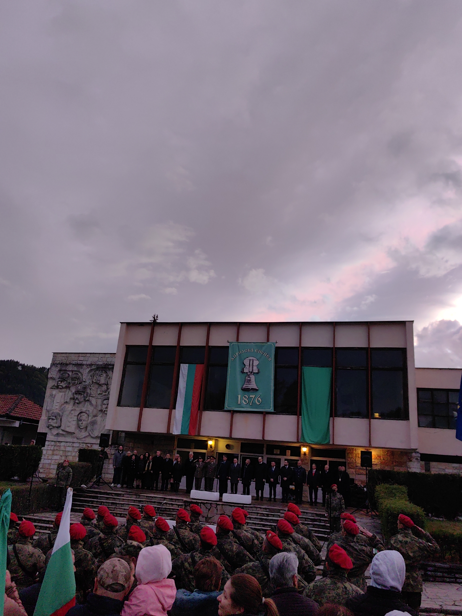 The image depicts an outdoor ceremony taking place in Klisura, Bulgaria, likely during dusk or early evening, as indicated by the dim lighting and cloudy, purplish sky.
The scene is set in front of a large, modern-style building. On the left side of the building's facade, there is a prominent bas-relief sculpture depicting several historical figures. Adorning the building are various banners: a vertical Bulgarian flag (white, green, and red stripes), a plain green banner, and a central green banner featuring a stylized bell, the year "1876," and the Cyrillic text "АПРИЛСКА ЕПОПЕЯ" (April Epic). These details strongly suggest the event commemorates the April Uprising of 1876.
In the midground, a group of officials and dignitaries, dressed in suits and coats, stands on a raised platform or steps in front of the building, observing the ceremony. Among them are also individuals in military or formal attire.
The foreground is filled with a crowd of spectators. Many people are seen from behind, including a child in a pink hoodie. A significant portion of the crowd consists of individuals in military-style camouflage uniforms with distinctive red berets. Some of these uniformed individuals are captured in the act of saluting, indicating their active participation in the formal proceedings. Small Bulgarian flags are also visible among the attendees.
The overall atmosphere is one of solemn commemoration and patriotism, highlighted by the historical references, military presence, and national flag displays.
