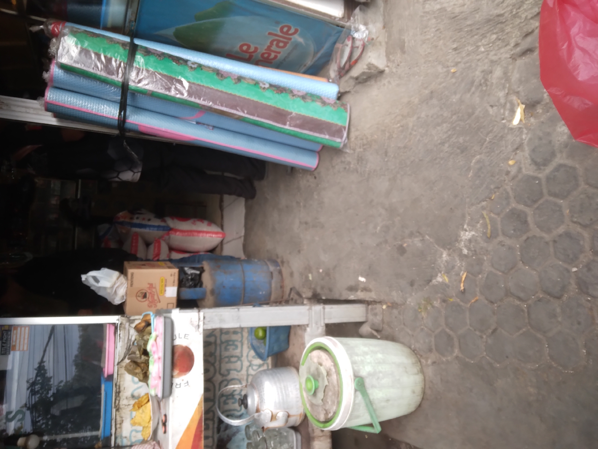 A street-level, medium shot depicts an outdoor food stall in Desa Pamijen, Kecamatan Sokaraja, Indonesia. In the foreground, a large, light green plastic bucket with a green lid and handle sits on a honeycomb-patterned paved surface. To the left, a silver metal teapot rests on a white counter alongside glass cups and a blue plastic container with a lime. Behind the counter, a selection of food items is displayed, including fried items on a white plate, and what appear to be wrapped snacks or rice cakes in a blue basket and on a grey tray. A cardboard box with an illustration of a person and text is also visible.
In the mid-ground, the stall's structure is evident, with a white shelf and metal supports. To the right, a stack of rolled-up items, possibly sleeping mats or fabric rolls, bound by black string, are propped against a blue cylindrical object, likely a gas tank. A blue tarp with red lettering, reading "Le Merale," is partially visible. Two individuals are present; one, wearing dark clothing, stands partially obscured behind the stall, while another, also in dark attire, is visible in the background. A white plastic bag is placed on the counter. The overall environment suggests a humid, possibly urban or semi-urban setting, with visible debris on the ground.
