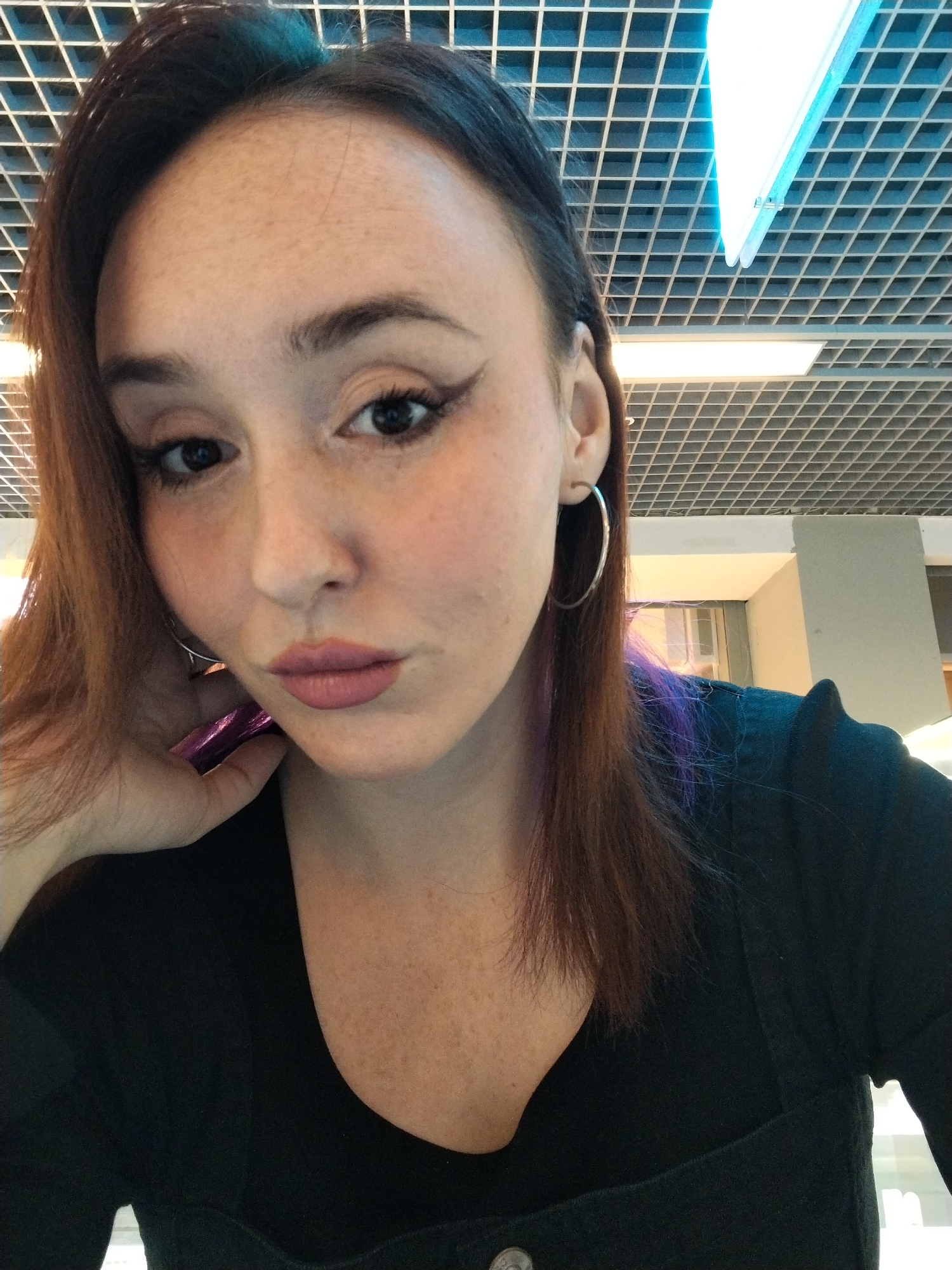A young woman is featured in a close-up, chest-up selfie, taken indoors. She has medium-length, reddish-brown hair with subtle purple highlights visible on the right side. Her face shows distinct features, including dark eyes enhanced with winged eyeliner, blush on her cheeks, and pink lipstick on her slightly parted lips, giving her a pensive or serene expression. Her skin has visible freckles across her forehead and cheeks. She is wearing large silver hoop earrings and a black top or dress with a square neckline. Her left hand is visible near her chin, holding a small, shiny purple object.
The background reveals an indoor environment, likely a public or commercial building in Izhevsk, Russia, characterized by a white grid-patterned suspended ceiling. Integrated into this ceiling are various artificial light sources. A prominent, elongated light fixture, emitting a cool blue light, extends diagonally across the upper right portion of the frame. Other warmer-toned, square-shaped lights are visible within the grid. Architectural elements, such as parts of walls or columns, can be discerned in the upper right. The lighting is bright and artificial, suggesting the photo was taken at any time of day under interior illumination. No text is visible in the image.