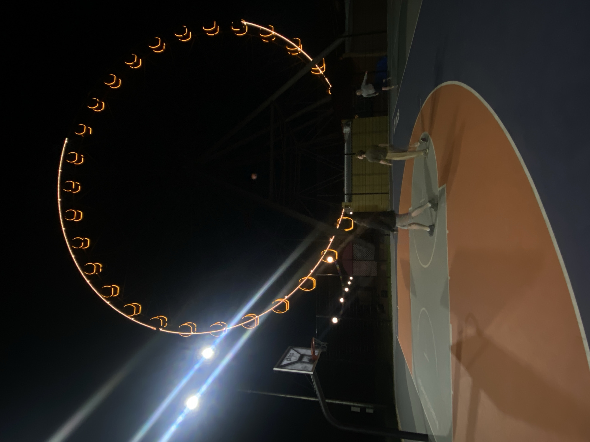 The image captures a nighttime scene at an outdoor recreational area, likely in Шебунино, Russia. On the right, a segment of a basketball court is visible, featuring a blue surface with an orange D-shaped key and white boundary lines. A basketball hoop with its backboard and net is positioned in the lower center. Three individuals are present on or near the court; one person stands closer to the hoop, while two others are further back. They appear to be casually standing, not actively playing.
In the background on the left, a large Ferris wheel dominates the view, illuminated by numerous warm orange lights that form a prominent arc against the dark night sky. Its structural frame can be faintly seen behind the cabins. Several bright artificial lights, possibly streetlights or court lights, are visible in the lower left, creating noticeable lens flares. The time of day is clearly night, as indicated by the dark sky and the reliance on artificial illumination. On the blue section of the basketball court, the word "DIVE" is faintly visible in white text. The overall atmosphere is lit by the glow of the Ferris wheel and the ground lights, creating a vibrant yet relaxed evening setting.