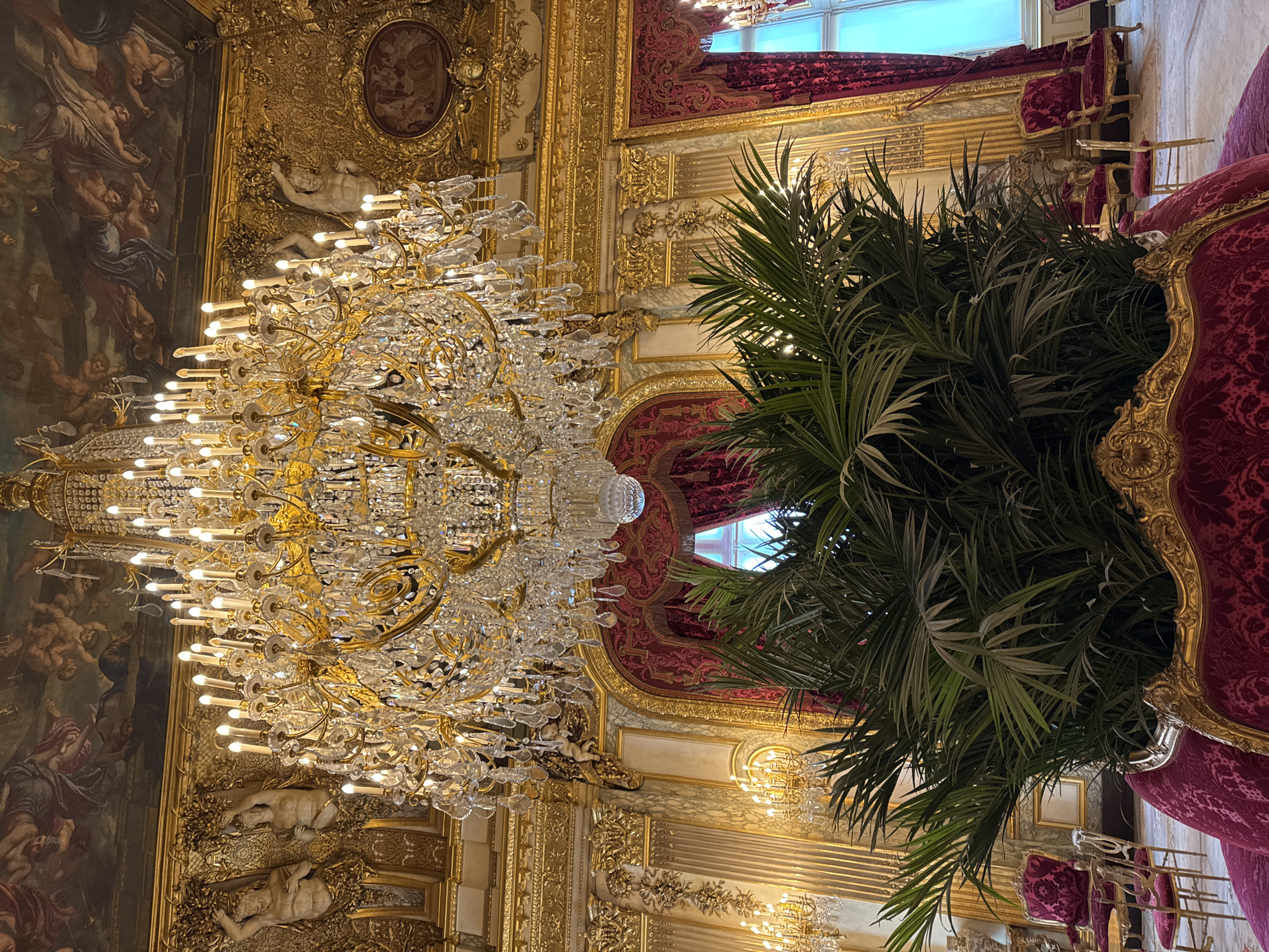 The image shows an interior view of a room within the Louvre Museum in Paris, France. The scene is dominated by an ornate, multi-tiered crystal chandelier hanging from the ceiling. The ceiling itself is highly decorated with frescoes and gilded moldings. The walls are paneled and ornamented with gold detailing, sculptures, and sconces. A large potted palm stands to the right of the chandelier, partially obscuring a window and the view beyond. Adjacent to the palm is an ornate red velvet chair with gold trim, part of a set of chairs.