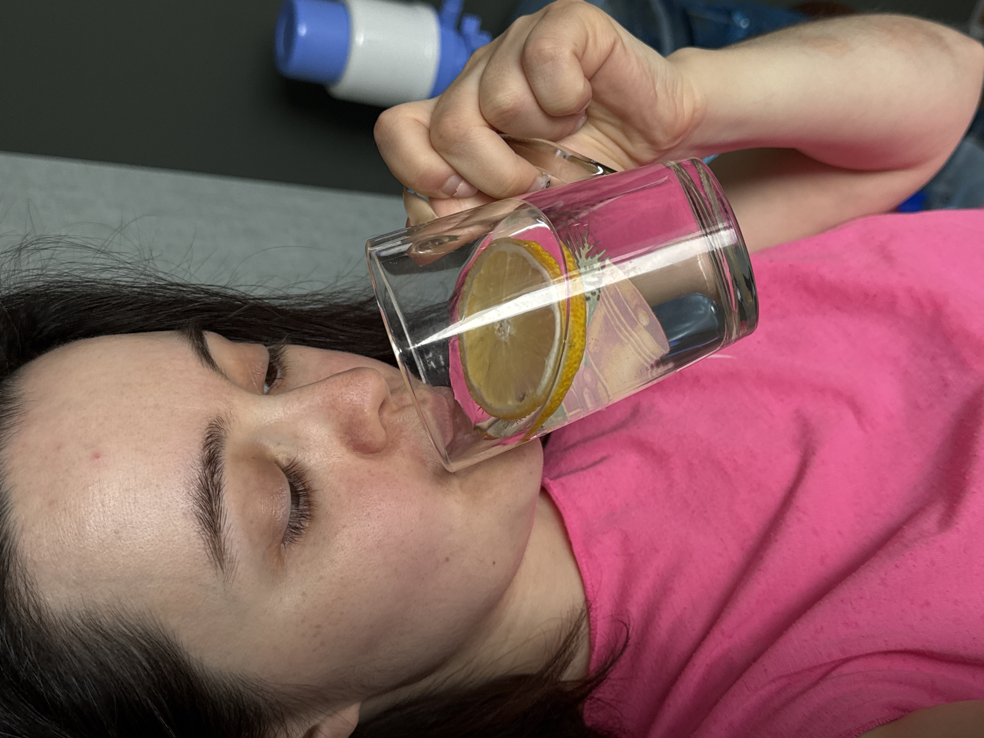 A woman with dark hair, wearing a pink shirt, is drinking from a clear glass of water with a lemon slice in it. The woman is lying down. In the background, there is a grey surface, blue jeans, and a white and blue object that resembles a water pump. There is no indication this image was taken in Izhevsk, Russia.