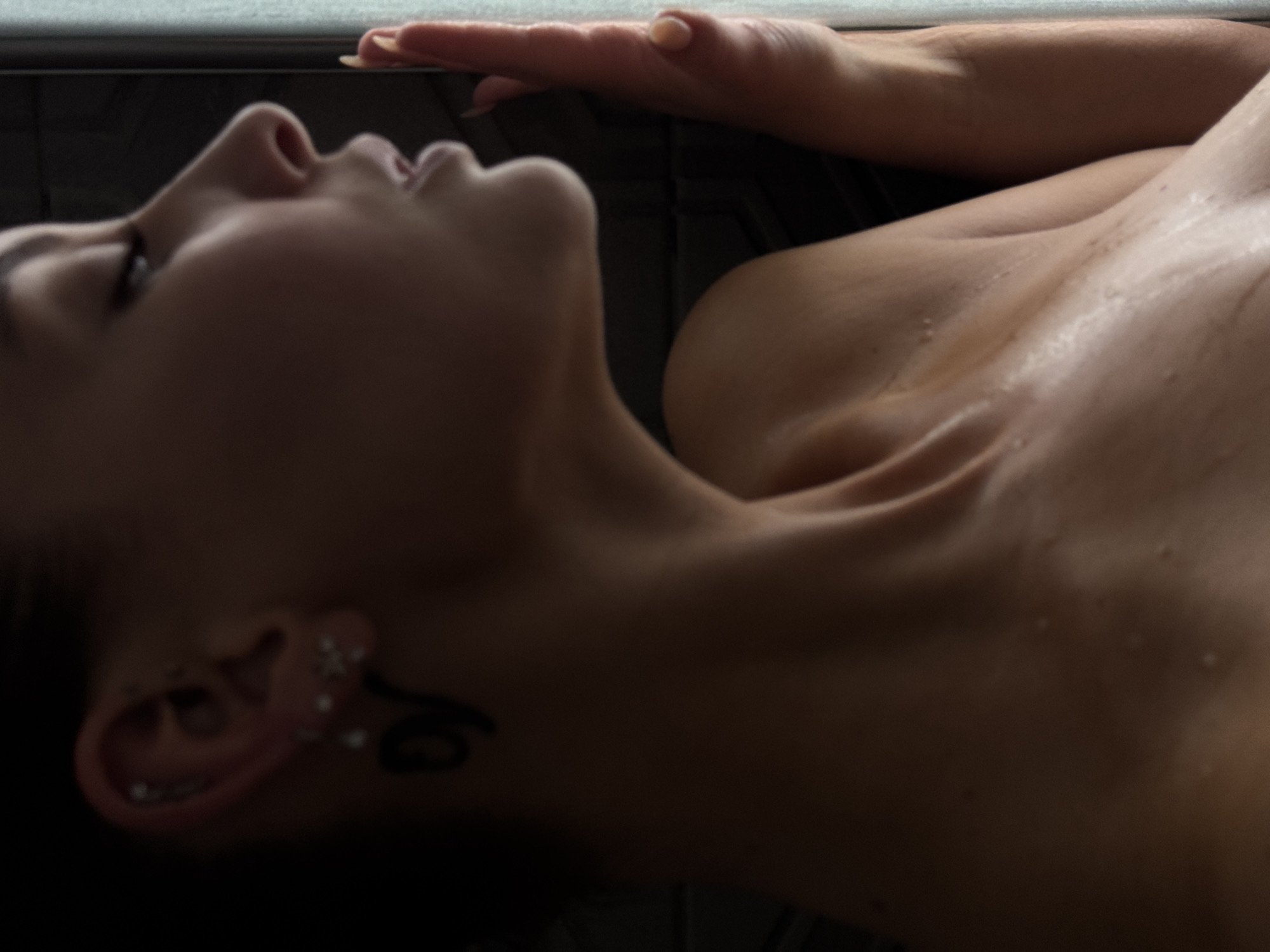 A woman's profile is captured in a dimly lit, intimate setting, possibly a shower, given the glistening water droplets on her skin. Her head is tilted slightly upward and to the left, with her eyes closed and lips parted as if in a moment of reflection or sensual experience. She has dark hair pulled back, revealing her ear adorned with multiple piercings and a dark, symbolic earring. A tattoo is visible on her upper chest, partially obscured by shadow. Her right hand is pressed against a textured, vertical surface, possibly a tiled wall or shower door. The lighting emphasizes the contours of her shoulder and collarbone, creating a dramatic chiaroscuro effect. The background is dark and indistinct, providing no specific location cues.