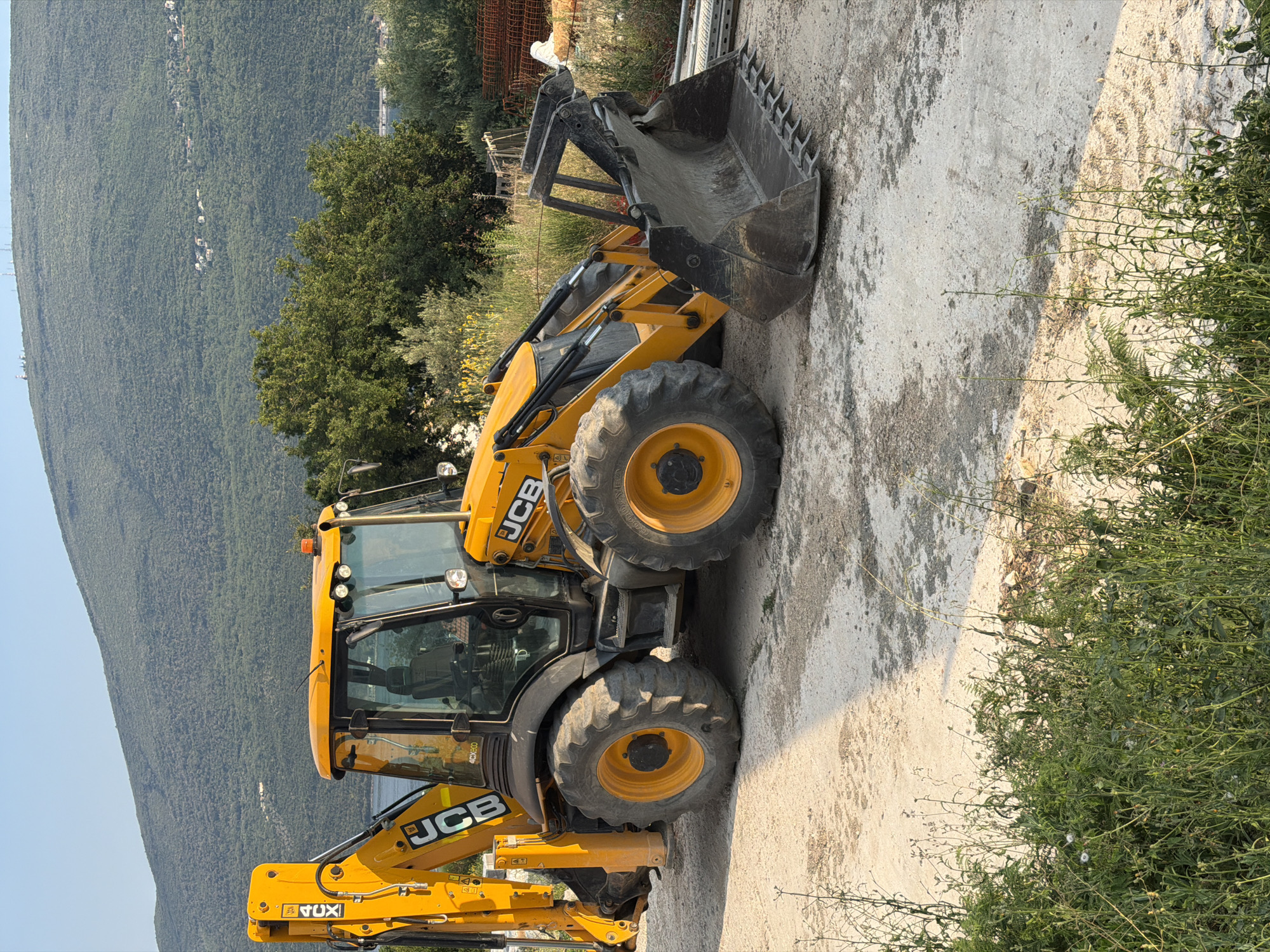A yellow JCB backhoe loader is parked on a dirt and gravel surface in Đenovići, Montenegro. The machine has a large front bucket and a backhoe arm. The cab is visible, with windows showing the interior. The tires are large and heavy-duty. In the background, there is a dense green hillside covered in trees, with some buildings scattered across it. The sky is clear and blue. Some construction materials, including rebar and metal scaffolding, are piled up to the right of the backhoe. Green vegetation and tall grass are growing in the foreground.