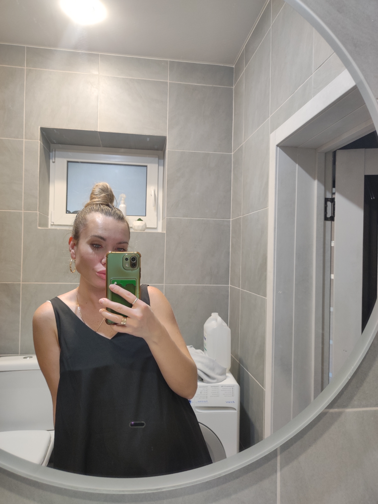 The image shows a selfie taken in a bathroom. The person taking the photo is a woman with her hair in a bun, wearing gold hoop earrings, a gold necklace, and a black top. She is holding a green phone in front of her face, obscuring part of her mouth. Behind her, there is a toilet to the left and a washing machine to the right, on which a white bottle and a folded grey cloth are placed. The bathroom is tiled with grey tiles. There is a window above the toilet with what appears to be small plant pots on the windowsill. To the right of the person is a doorway with a partially open door. The location in Votkinsk, Russia, cannot be determined from the given information.
