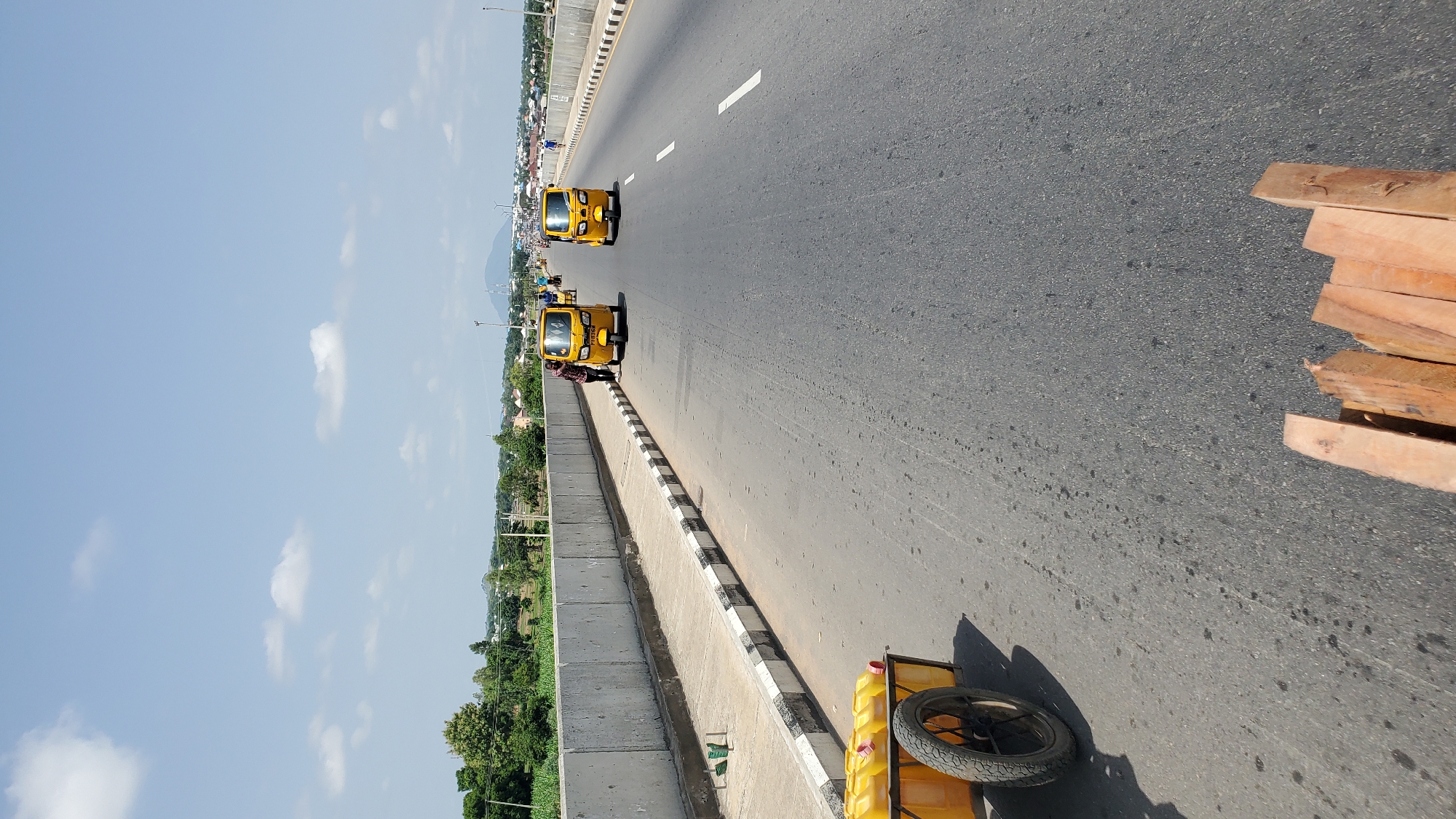This media file captures a view of a highway in Jalingo, Nigeria. The scene features two yellow auto-rickshaws, also known as Keke Napep, driving on the road. One auto-rickshaw is in the foreground, and another is further ahead. A person in a checkered shirt is leaning on the side of the road near the foreground auto-rickshaw. To the right of the road, a stack of wooden planks is visible. In the foreground, a yellow cart with several yellow jerrycans and a spare tire is parked.
The setting is a multi-lane highway with a concrete barrier separating the road from a lower level. Beyond the barrier, there is greenery and buildings, suggesting a semi-urban or rural fringe. The sky is blue with scattered white clouds, indicating daytime with fair weather. The sunlight casts shadows on the road, suggesting the time of day is likely morning or afternoon. The overall impression is of a typical day on a Nigerian highway, with local transportation and roadside activity. No specific text is clearly legible.
