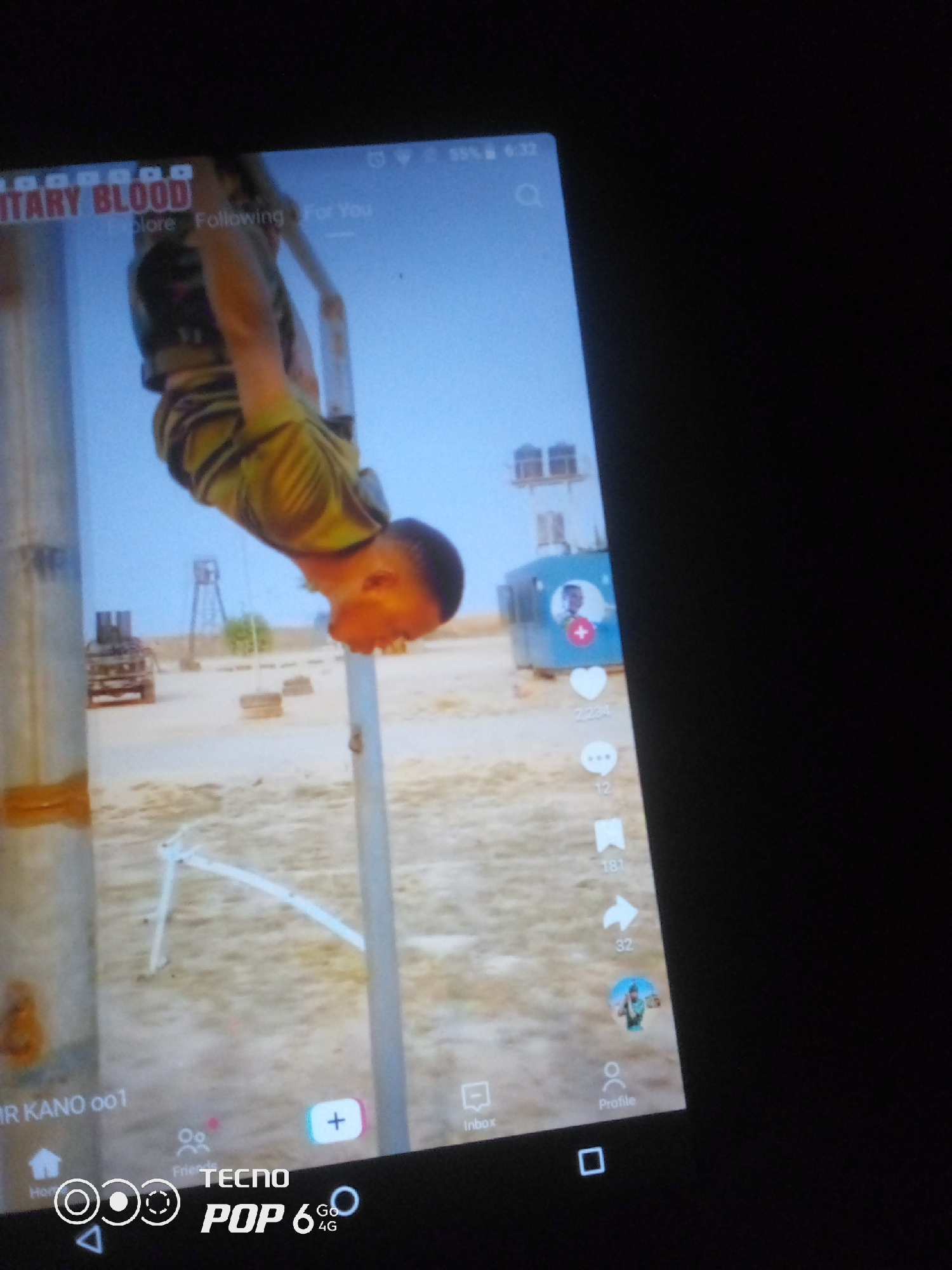 A video is displayed on a tablet screen, showing a person hanging upside down from a horizontal bar. The person is wearing a camouflage uniform. In the background, there are structures that appear to be military in nature, including a vehicle and elevated platforms with shelters. The ground is sandy and appears to be an outdoor training area. On the right side of the screen, there are social media icons, including a heart, speech bubble, bookmark, and a profile icon. The bottom of the screen shows navigation icons and the "TECNO POP 6 4G" branding. The video appears to be set in Howayi, Nigeria, as indicated by the text "MR KANO 001" visible on the screen.