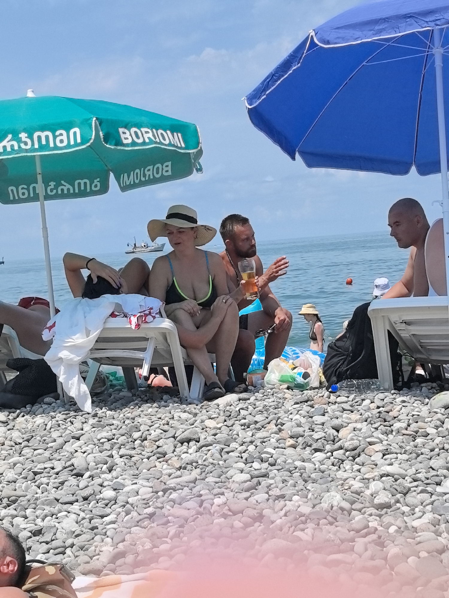 The image depicts a sunny day at a pebble beach in Batumi, Georgia, with several people relaxing under beach umbrellas. In the foreground, two individuals are seated on white plastic lounge chairs. One person, wearing a wide-brimmed straw hat and a dark bikini top with neon green straps, is looking towards the right. The other person, lying on a lounge chair, has their head resting on their hand and a white cloth draped over them.
Behind them, a man with a beard is holding a glass of amber liquid and gesturing with his other hand, appearing to be in conversation with another man who is facing him. This second man, with a shaved head, is also seated on a lounge chair and looking towards the man with the beard.
In the background, the sea is visible under a blue sky with scattered clouds. A small boat is sailing in the distance. The beach is covered in pebbles. Several plastic bags containing various items, possibly drinks and snacks, are on the ground near the people. A small child in a straw hat and bikini is visible in the water. The presence of beach umbrellas with Georgian text, specifically "Borjomi," strongly suggests the location is along the Batumi coastline.