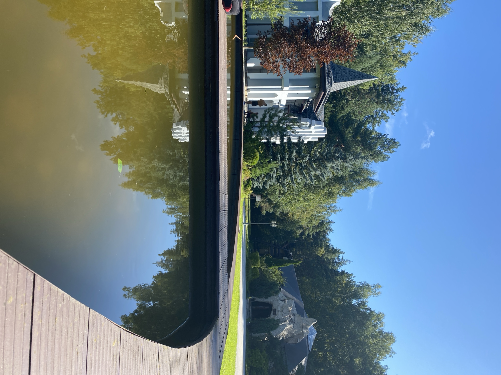 The image depicts an outdoor scene with a focus on a body of water and surrounding architecture. In the foreground, a wooden deck borders a murky green body of water that reflects the sky and surrounding environment. The water's surface shows a distorted reflection of a white building with a pointed turret and a dark-roofed structure resembling a cave entrance with a glass-paneled roof.
Behind the water and deck, the white building features a distinct pointed turret topped with dark grey material. It has multiple arched doorways and windows, and a red-leafed tree stands to its left. Further to the right, the cave-like structure is partially visible, blending into the natural landscape.
The background is dominated by a dense forest of green trees under a clear blue sky with a few wispy clouds. The overall setting appears to be a park or resort area. Two individuals are visible standing near the white building on a paved pathway. One is a man standing upright, and the other is seated on a bench. The environment suggests a tranquil, possibly recreational, location. The image does not contain any information to indicate the specific location within the city of Chulis, Russia.