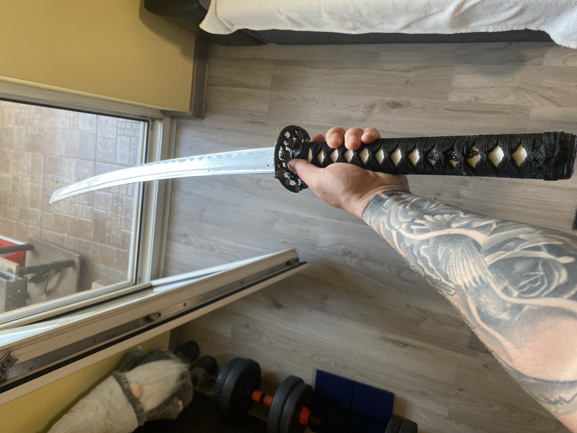 The image captures a first-person view of a human right arm holding a katana, situated indoors in Varna, Bulgaria. The arm is heavily tattooed with greywash artistry, depicting what appears to be a large bird and floral elements. The hand firmly grasps the katana's handle (tsuka), which is wrapped in black cord over a white material, forming a traditional diamond pattern. The metallic blade is visible above a dark, openwork handguard (tsuba). The indoor environment features light-toned laminate wood flooring and solid yellow-painted walls. To the left, a large grey fabric gnome decoration with a white beard is positioned on the floor, next to a stack of black dumbbells with red connectors resting on a blue mat. A white-framed glass sliding door or window, partially ajar, reveals an outdoor space paved with terracotta-colored square tiles. On the right side of the frame, a section of furniture, possibly a bed or sofa, is covered with a white, textured throw blanket.
