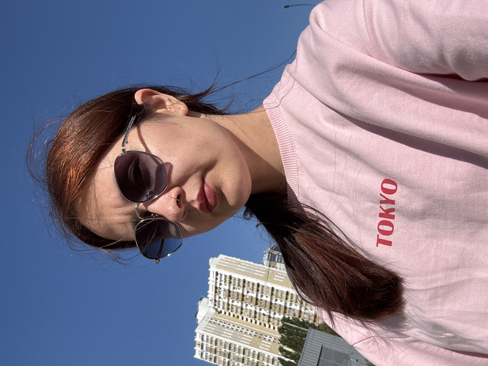 A young woman with auburn hair, wearing sunglasses and a pink sweatshirt with "TOKYO" printed in red, poses for a selfie outdoors under a clear blue sky. In the background, a tall, multi-story residential building is visible. The scene appears to be in a city, possibly Naberezhnye Chelny, Russia, given the architectural style of the building and the overall urban environment. The lighting suggests it is daytime.