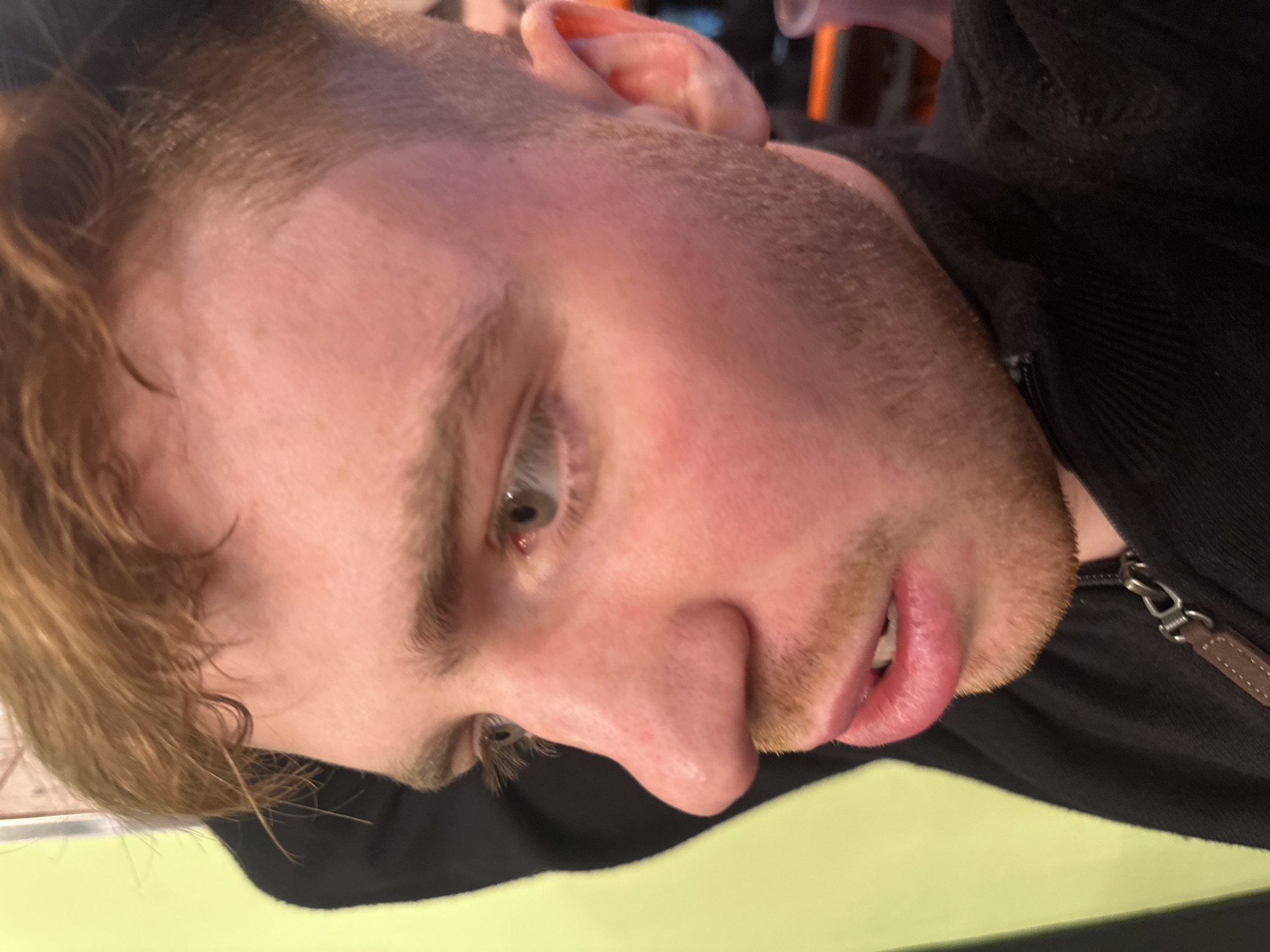 A close-up, profile image depicts the left side of a fair-skinned male subject with reddish-blonde hair and light green eyes. The subject has reddish-blonde stubble or a short beard. His face appears flushed, particularly on the cheeks and nose. The mouth is slightly open, revealing teeth and the pink interior of the lips. He wears a dark, possibly black, zip-up garment featuring a metal zipper and a brown, leather-like pull tab. The subject's left arm is partially visible behind his head, obscured by hair and clothing. The background includes a solid lime-green surface on the left. Blurred elements are visible behind the subject's head on the right, including an orange-lit area and a translucent purple object. The specific location within New York City, United States of America, cannot be determined from the image.