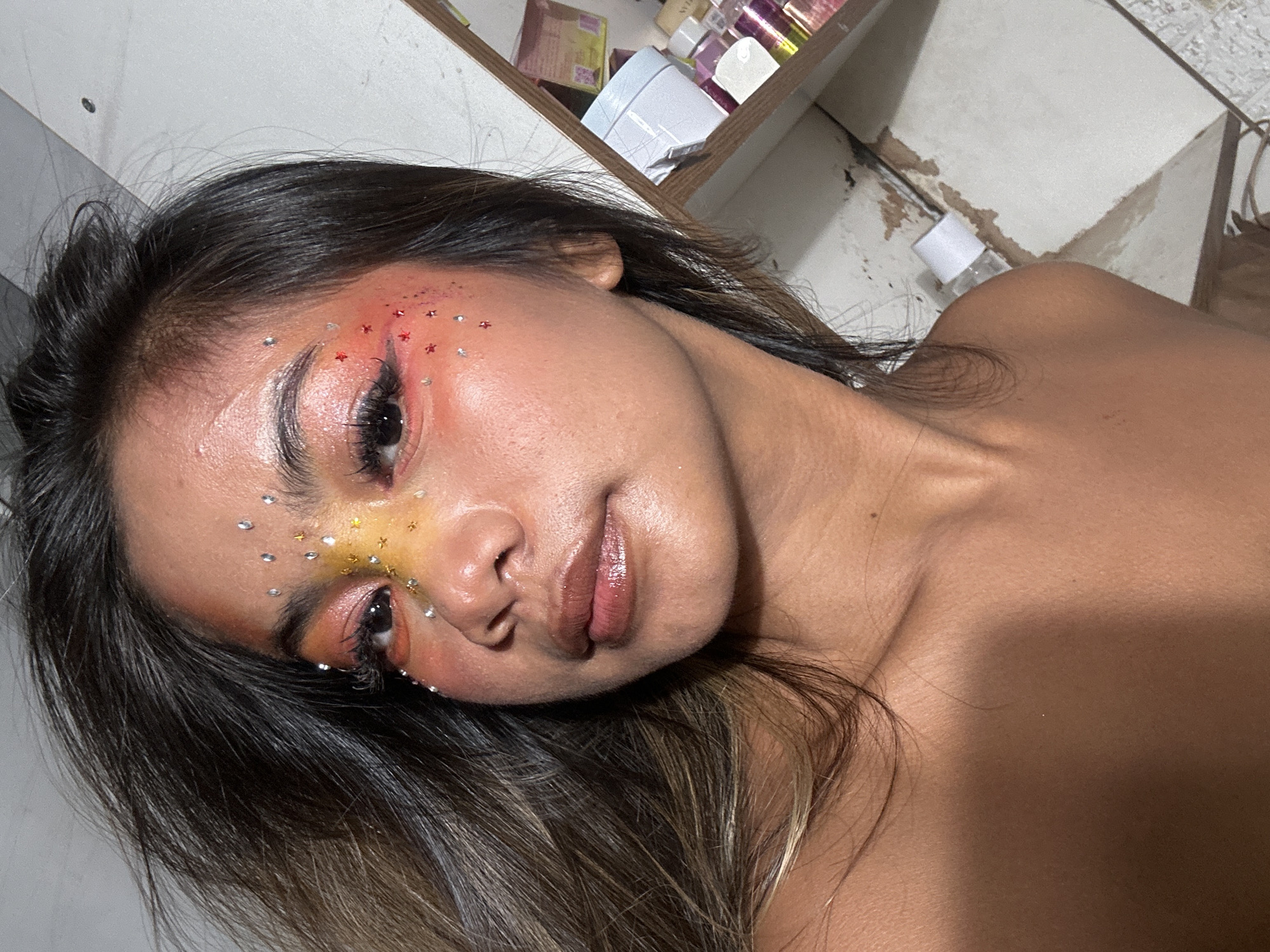 A young adult female with long, dark hair with brown highlights is shown from the chest up. She has tan skin and her face is adorned with a variety of colorful makeup, including red, orange, and yellow eyeshadow, as well as small, iridescent gemstones in a clustered pattern around her eyes and forehead. Her lips are glossy with a nude color. The background includes a partial view of a shelf with assorted bottles and containers, as well as a wall and floor with some damage. No specific location information in North Jakarta, Indonesia is discernible from the image.