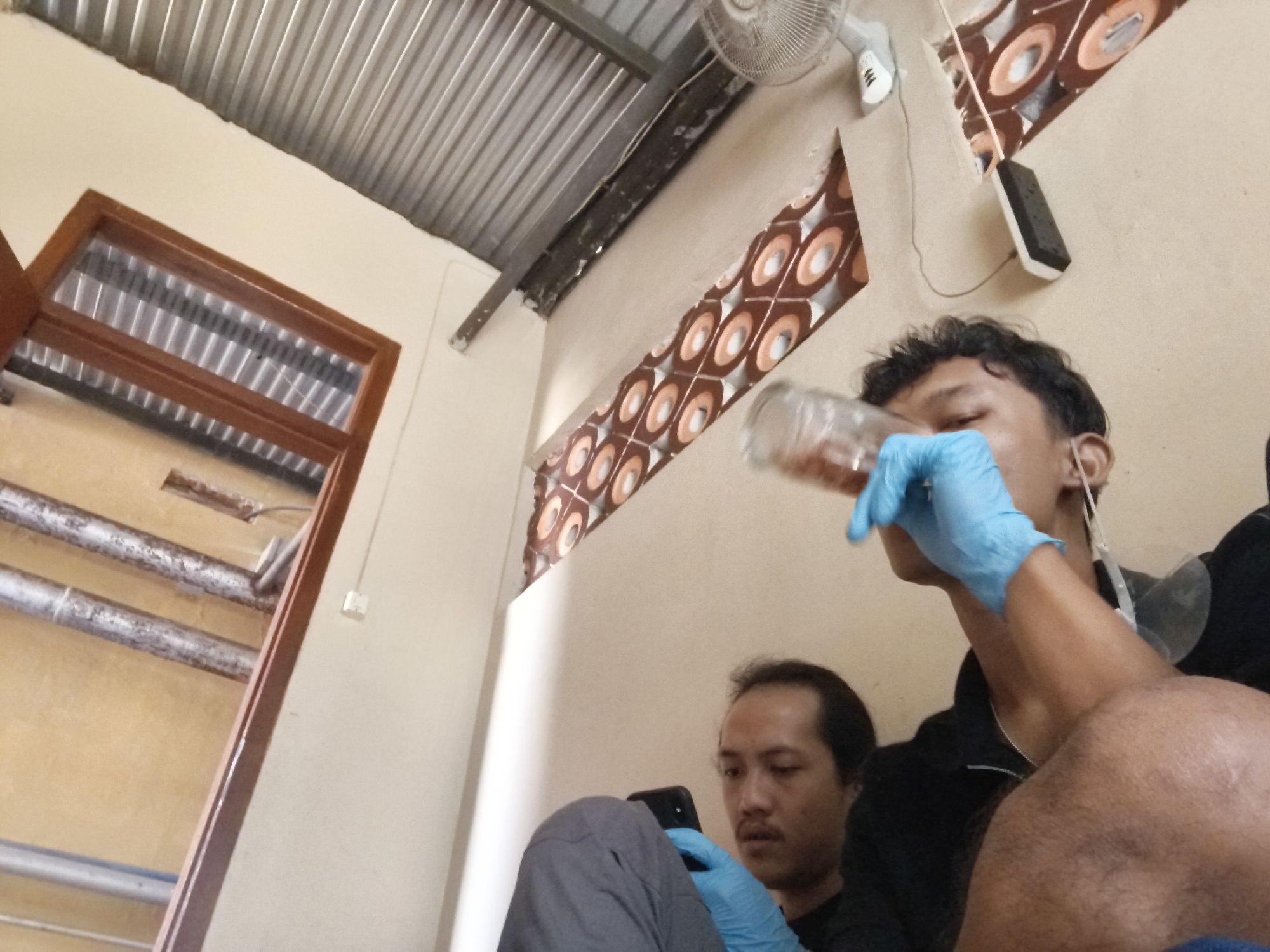 A low-angle, wide shot captures a dimly lit interior scene, possibly a room in Pekalongan, Indonesia, with corrugated metal roofing visible at the top. Two men are present. In the foreground and to the right, a man wearing a blue glove is holding a glass bottle to his mouth, suggesting he is drinking. He has dark hair and is wearing a black top. Slightly behind and to the left, a second man, also wearing a blue glove, is looking down at a black smartphone in his hands. He has dark hair and a mustache, and is wearing gray pants and a dark top. The room features light-colored walls with a decorative patterned opening and a wooden-framed doorway on the left. Pipes are visible running along the ceiling. A ceiling fan is mounted near the metal roof.