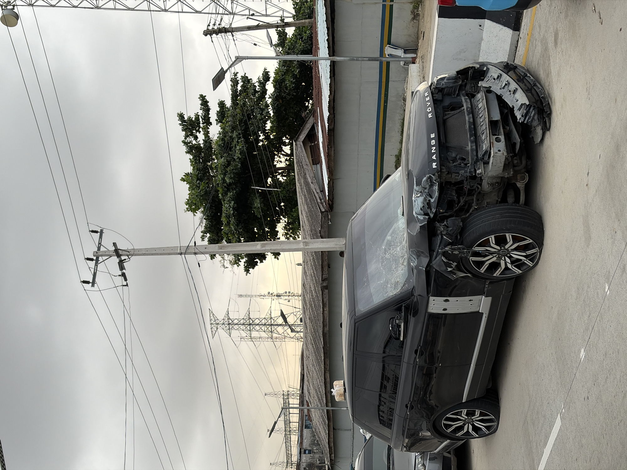 In Igboefon, Nigeria, a black Range Rover is depicted with extensive front-end damage, suggesting a recent collision. The vehicle's hood is crumpled and displaced, and the front bumper is detached and hanging low. The headlights are damaged, and the radiator is exposed. One of the front tires appears flat. The Range Rover is positioned on a paved road with a white line marking. In the background, high-voltage power lines and transmission towers are visible, along with some buildings and trees. The sky is overcast. A loaf of bread is precariously placed on the roof of the damaged vehicle.